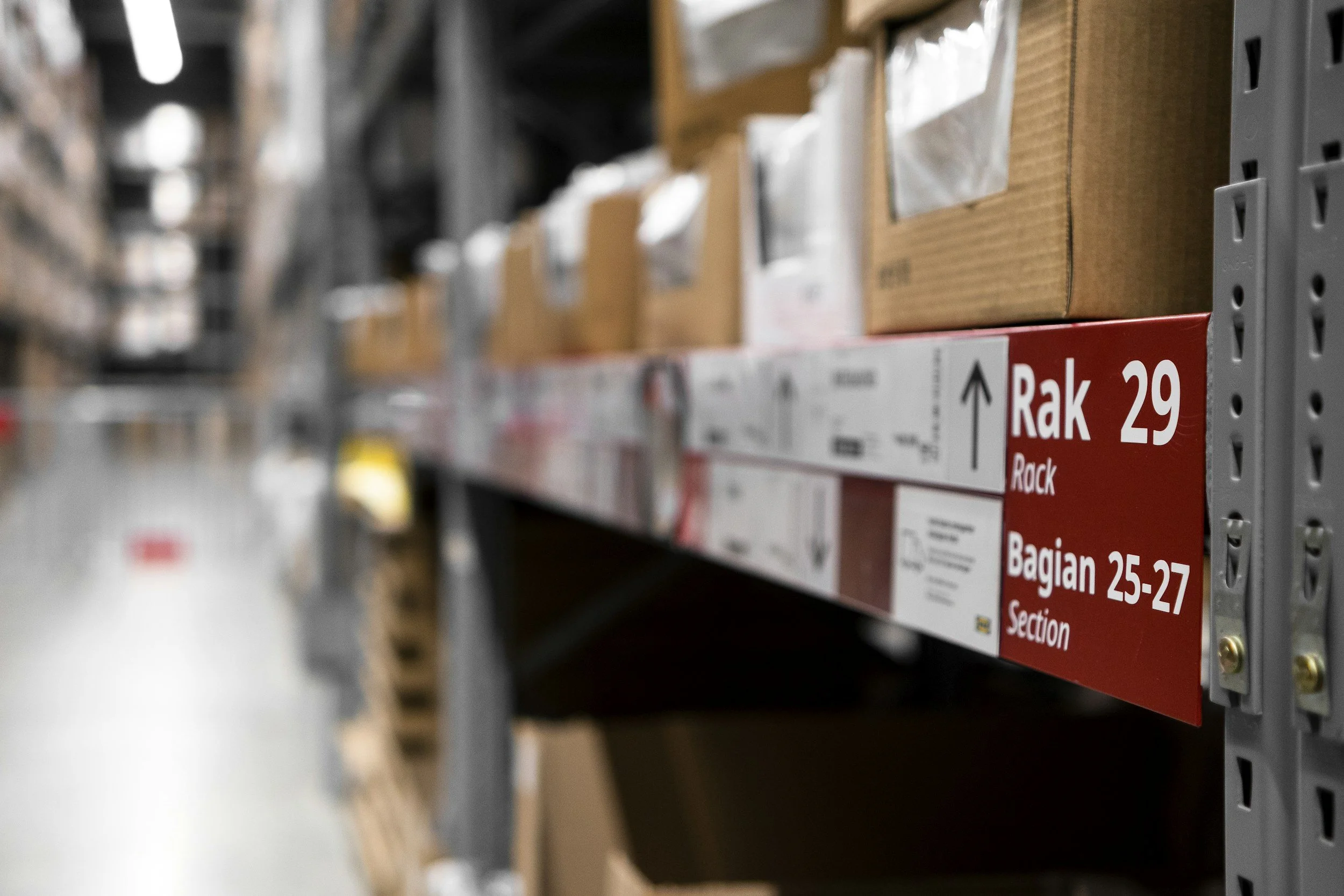 Warehouse aisle with metal shelving holding cardboard boxes and packaging materials, with a sign indicating section Rak 29, Bagian 25-27.