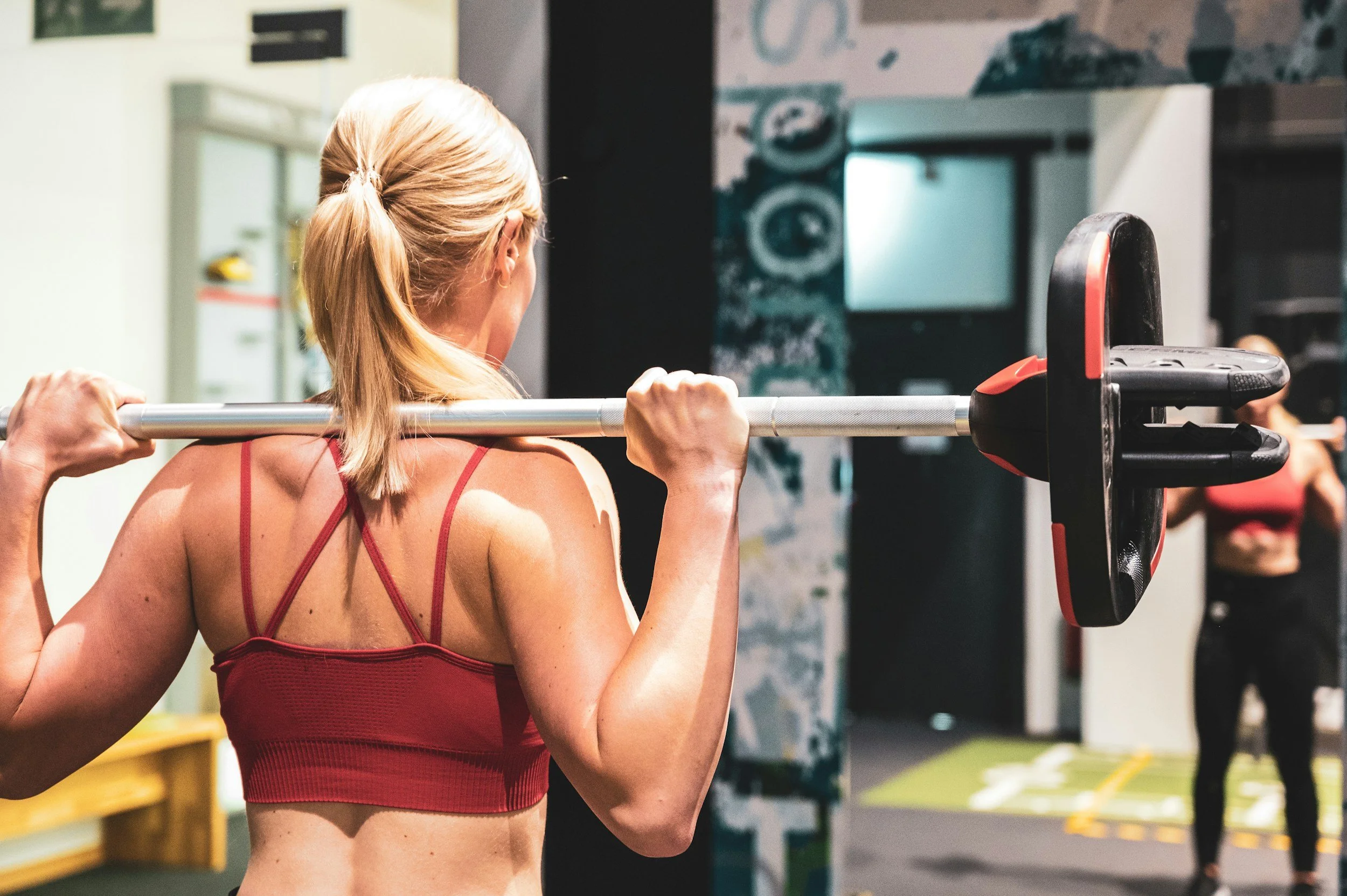 A woman with blonde hair in a ponytail lifting a barbell while standing in a gym, with another woman in workout clothes in the background.