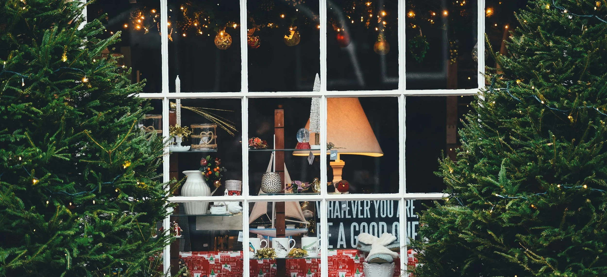 View through a window showing Christmas decorations inside a cozy room, with two decorated Christmas trees outside.
