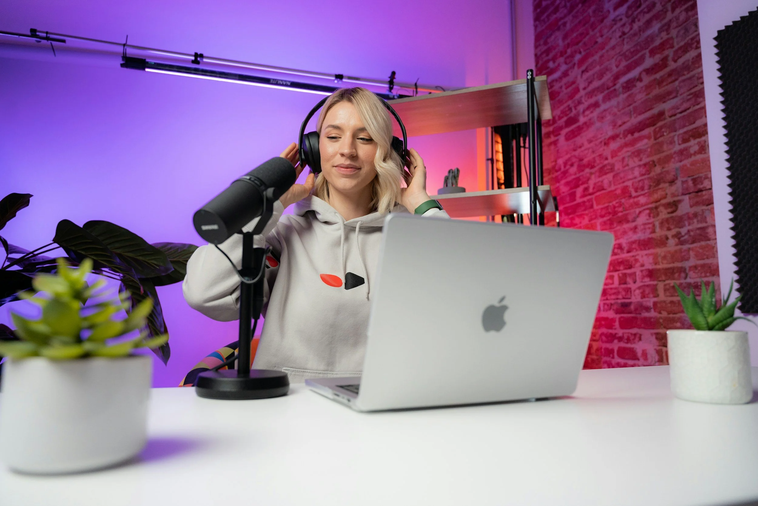A woman with blonde hair wearing headphones sitting in front of a microphone and laptop in a colorful recording studio with plants and brick walls.