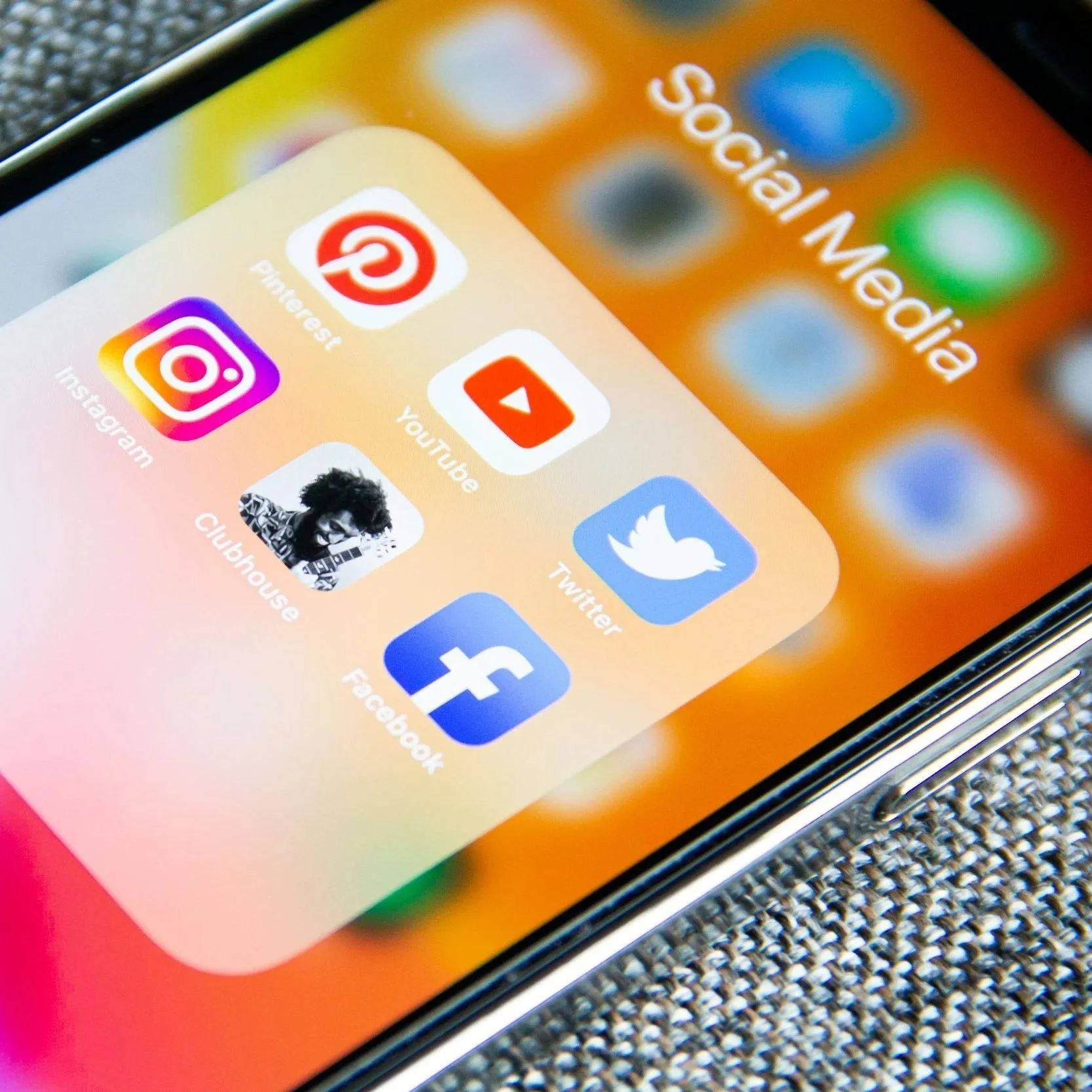 Close-up of a smartphone screen showing social media app icons in a folder labeled 'Social Media', including Pinterest, YouTube, Instagram, Twitter, Facebook, and Clubhouse, with a textured surface underneath.