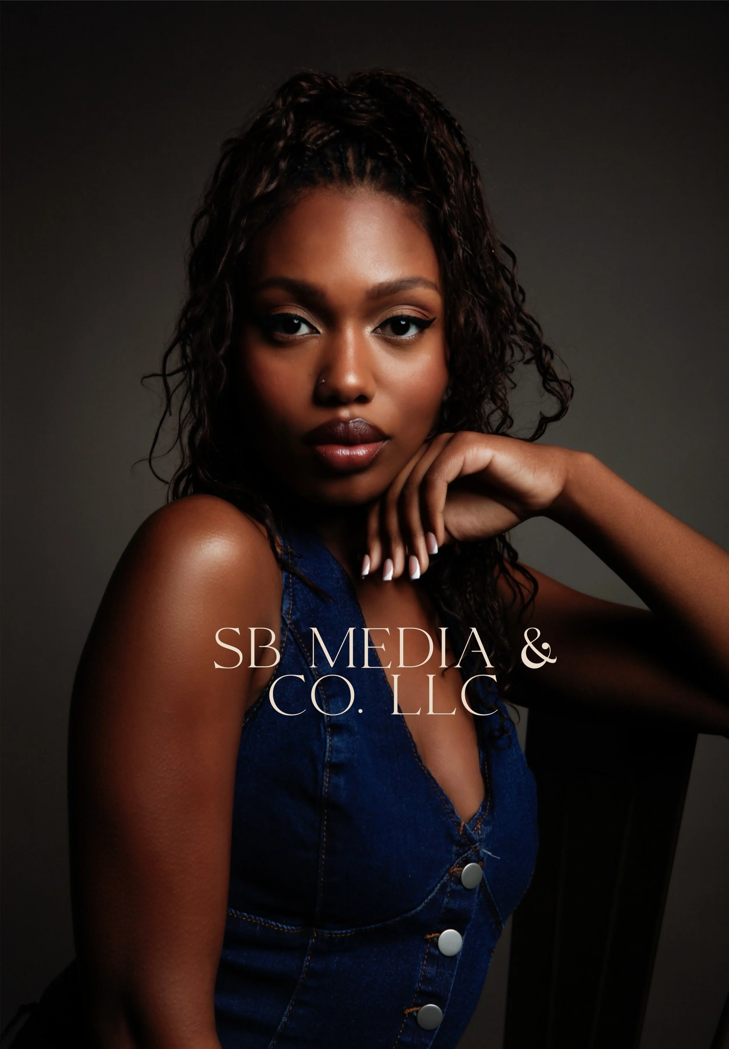 Portrait of a young woman with dark curly hair, wearing a denim top, against a dark background.