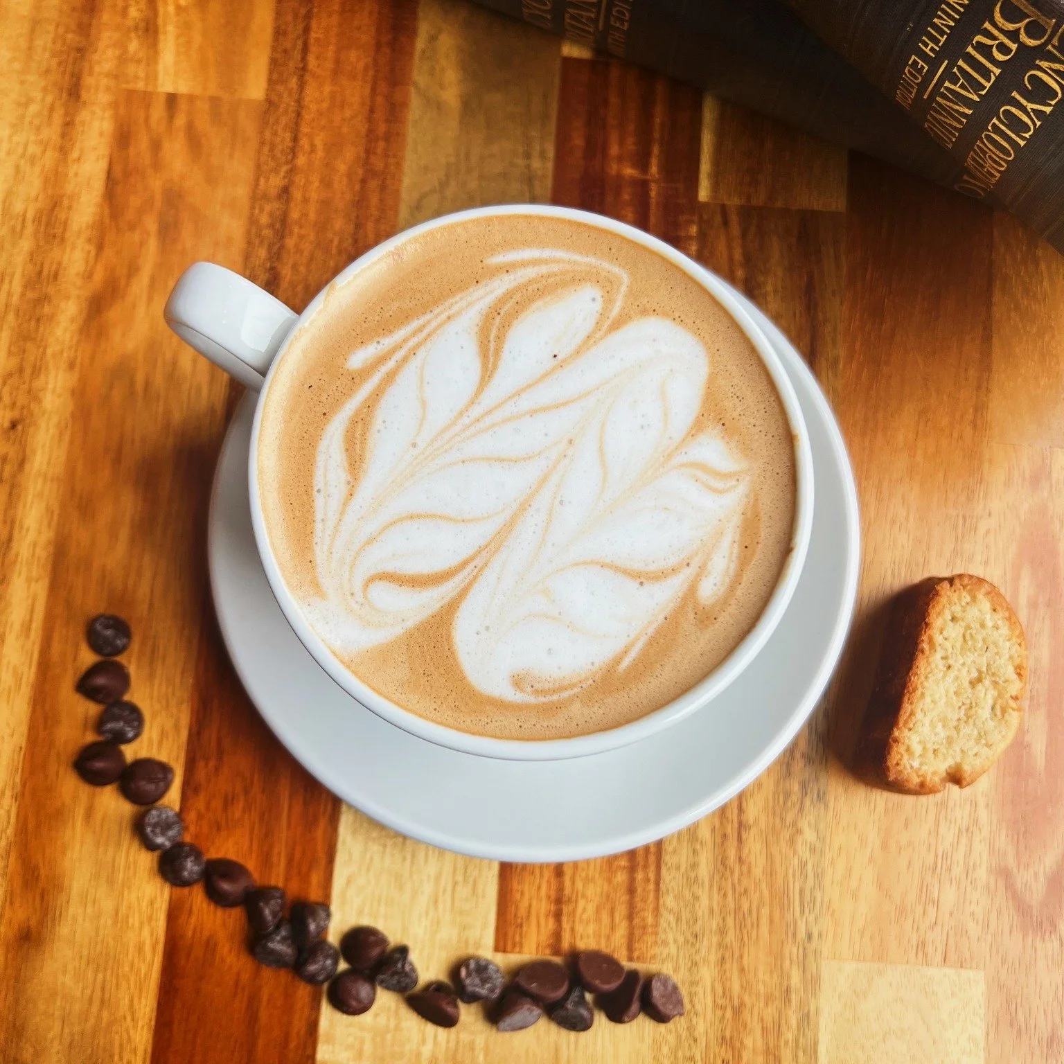A cup of coffee with latte art on top, placed on a white saucer, on a wooden table. Next to the cup are coffee beans and a biscotti.