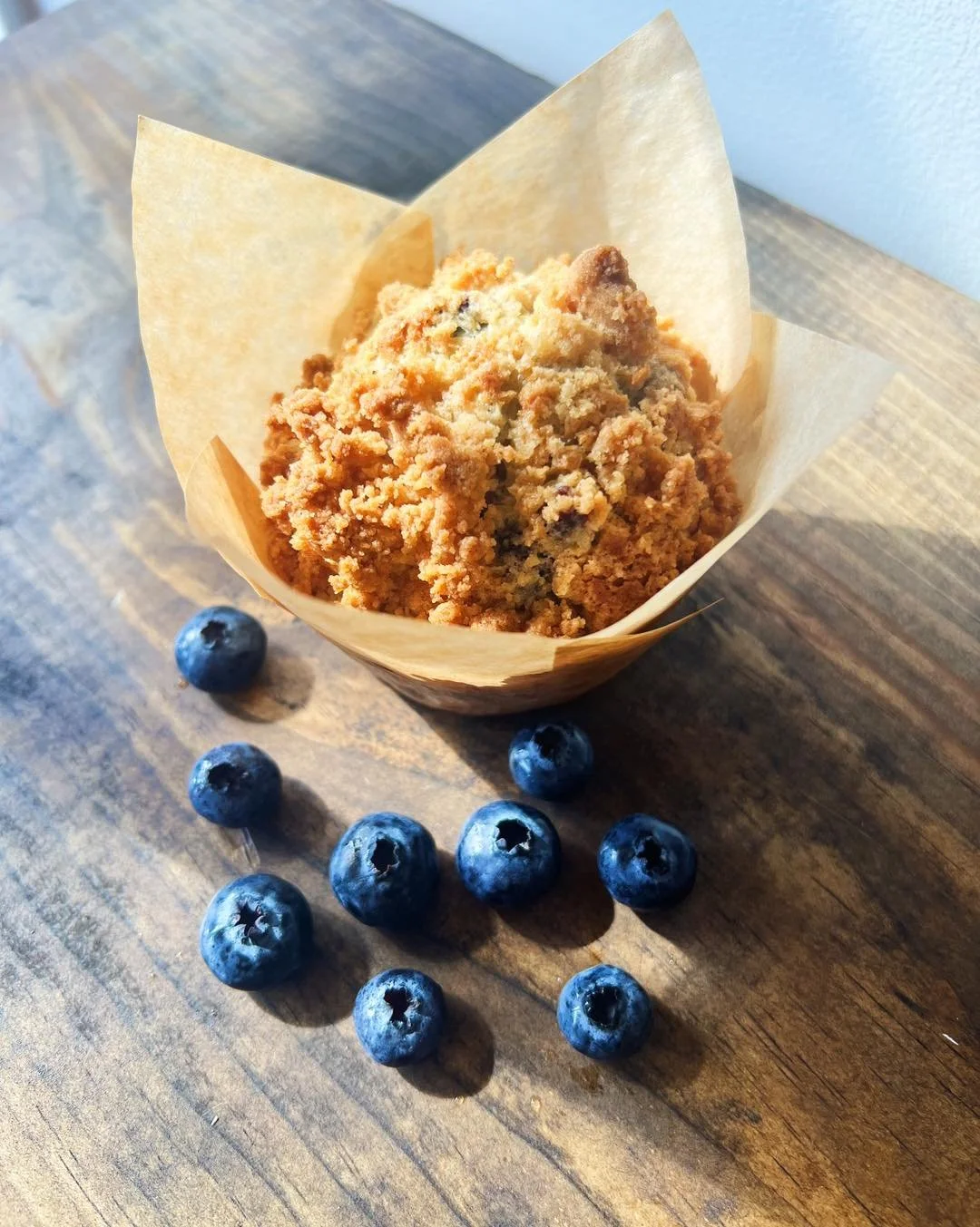 A blueberry muffin in parchment paper on a wooden surface surrounded by fresh blueberries.