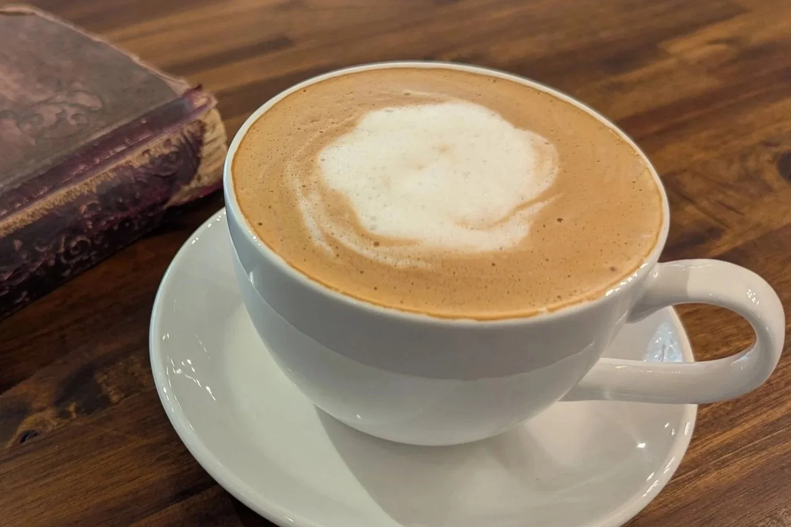A white ceramic cup filled with cappuccino topped with foam, sitting on a saucer on a wooden table, with a chocolate pastry in the background.