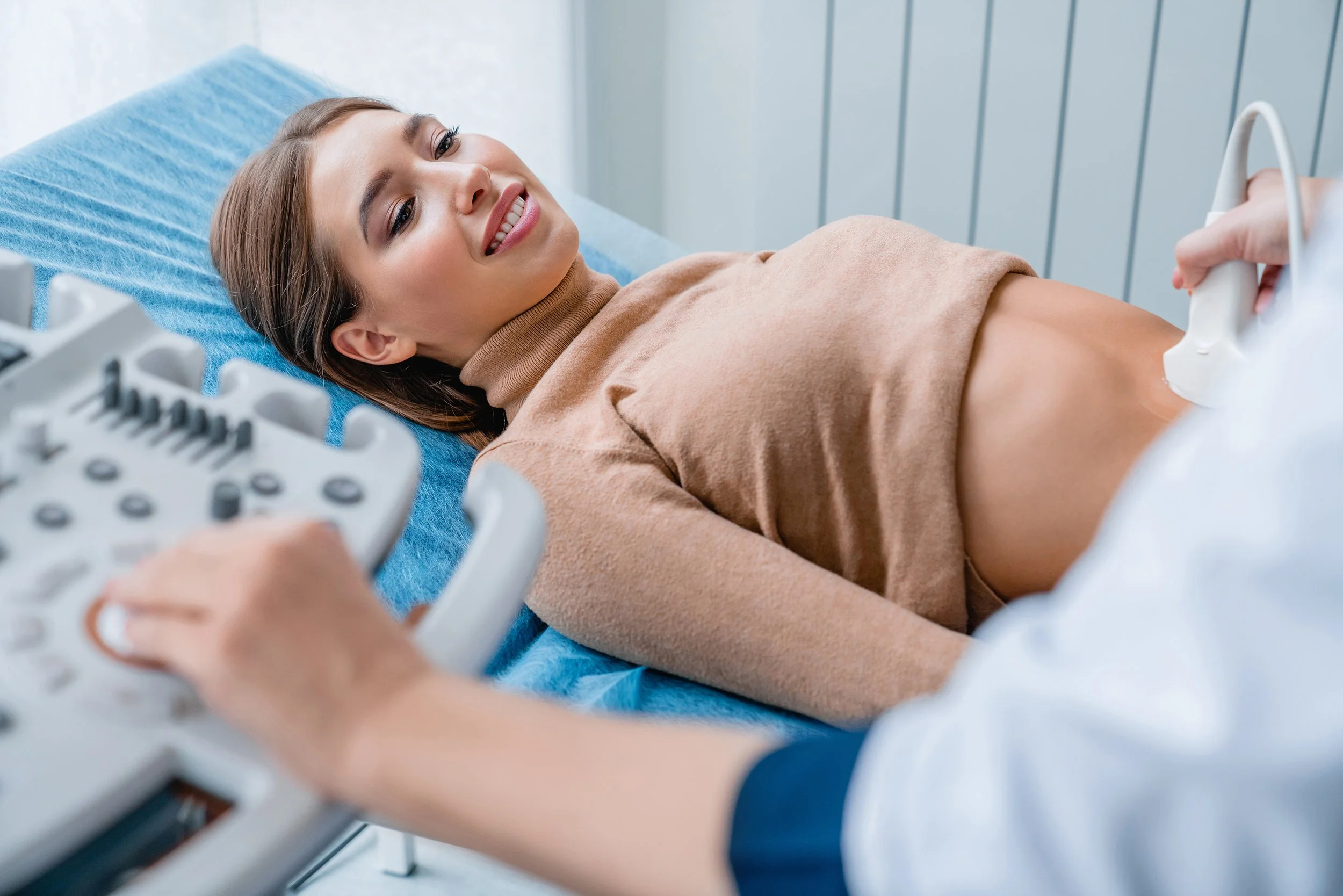 Woman lying on a medical examination table receiving an ultrasound scan, with medical equipment visible in the foreground.