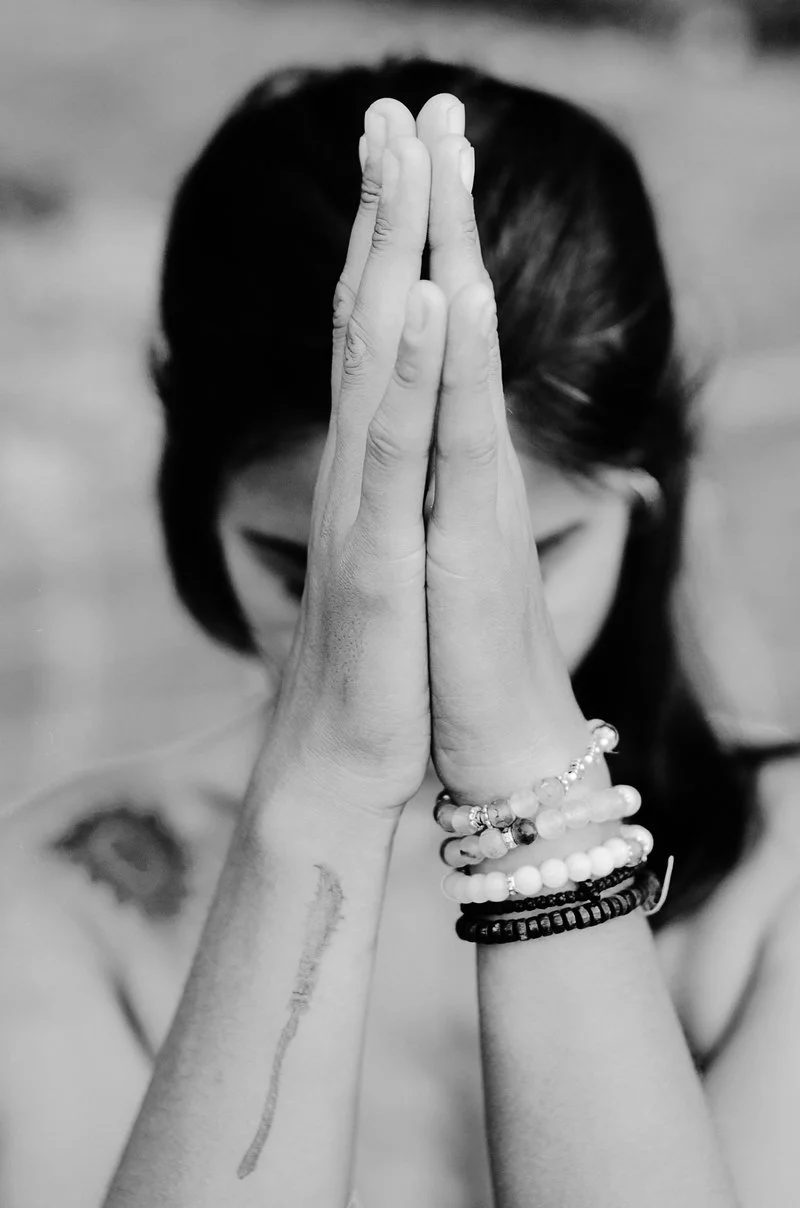 A woman with dark hair held back, raising her hands in prayer or meditation with palms together, wearing multiple beaded bracelets on her wrists.