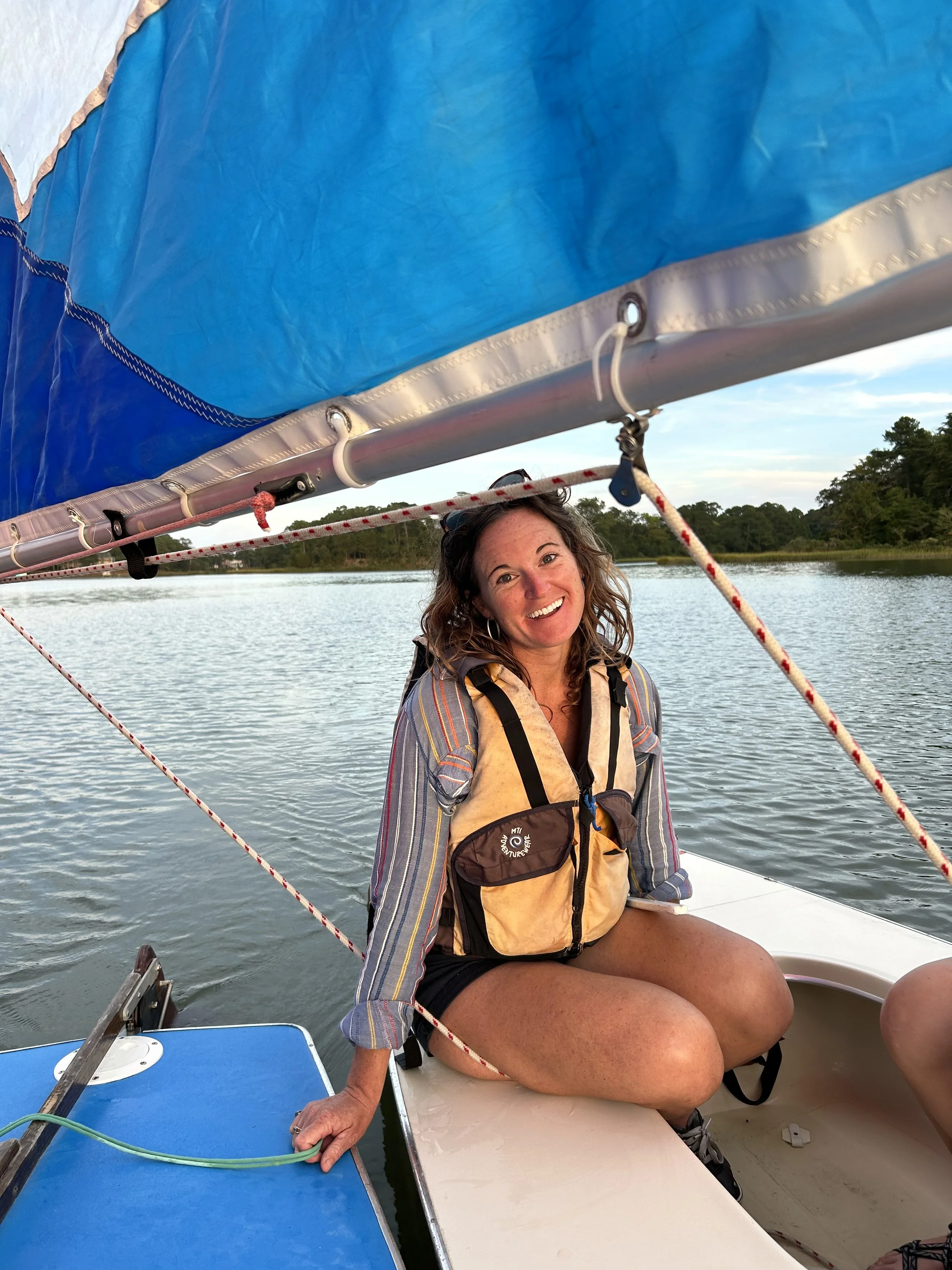 A woman sitting on the edge of a boat, smiling, wearing a life jacket, with a body of water and trees in the background.