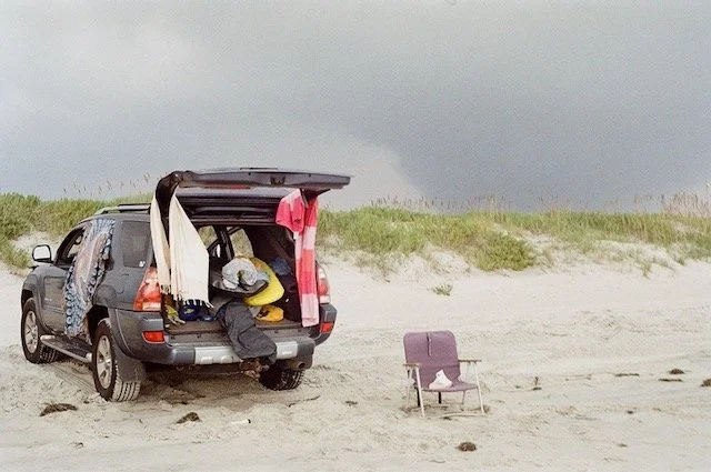 A person preparing to surf or swim, sitting behind a gray SUV with an open trunk, on a sandy beach with some grass and cloudy sky in the background, and a purple beach chair nearby.