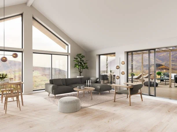 Modern living room with large windows, gray sectional sofa, round coffee table, potted plant, and sliding glass doors leading to a balcony with mountain view.