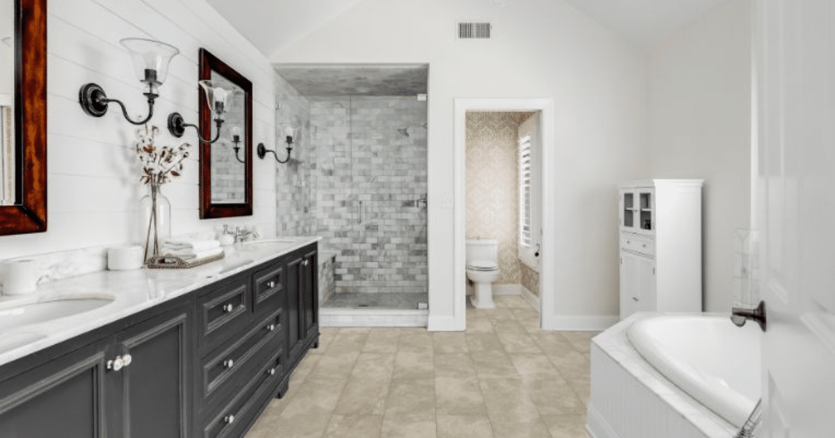 A spacious bathroom with a large dark gray vanity, white countertop, and two sinks. There are two mirrors above the vanity with wall-mounted light fixtures. The shower has gray stone tiles, and a separate toilet room is visible. There is a bathtub on the right side, and the floor is tiled with beige tiles.