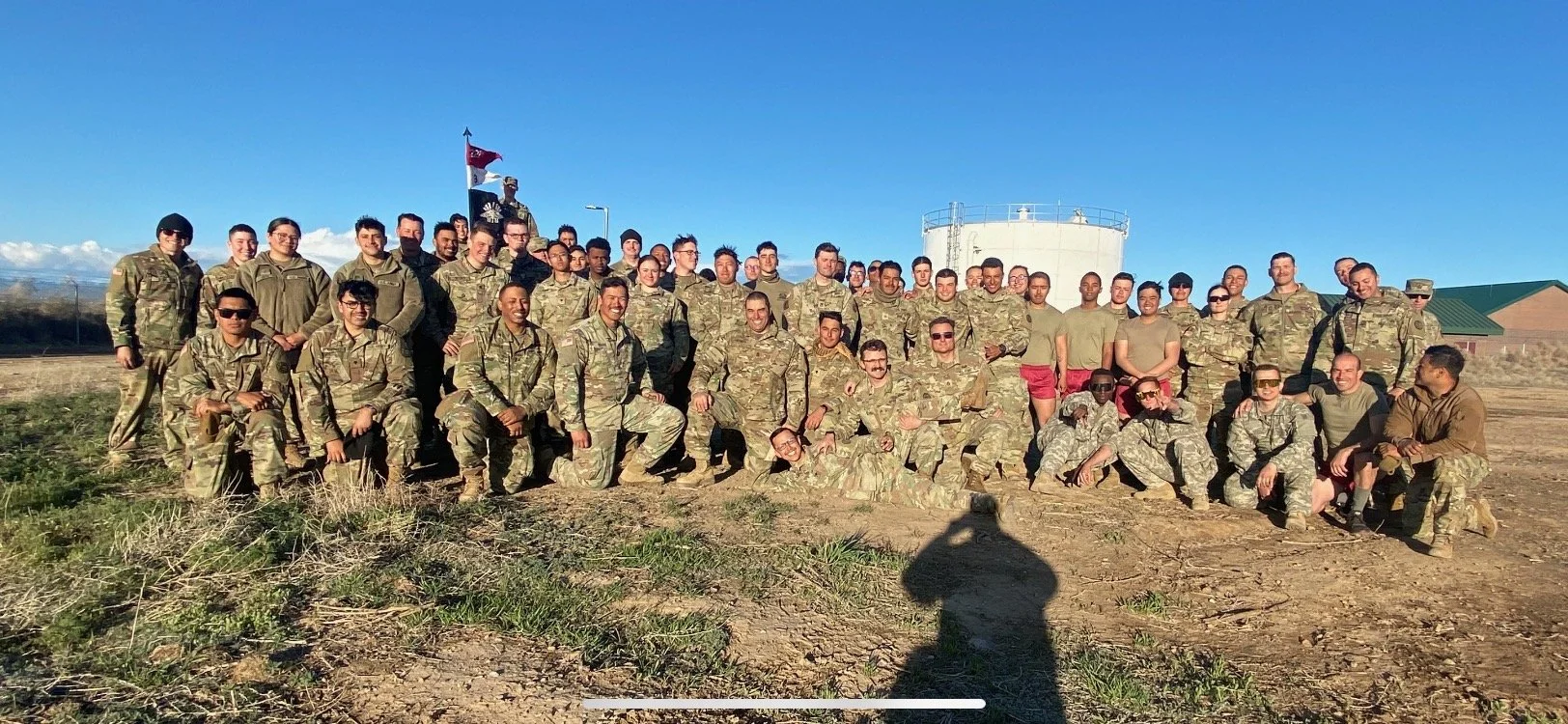 Large group of soldiers in camouflage uniforms led by elect judicial candidate 8th judicial district court department 28 alex quagge posing outdoors on a dirt field with a clear blue sky and water reservoir in the background.