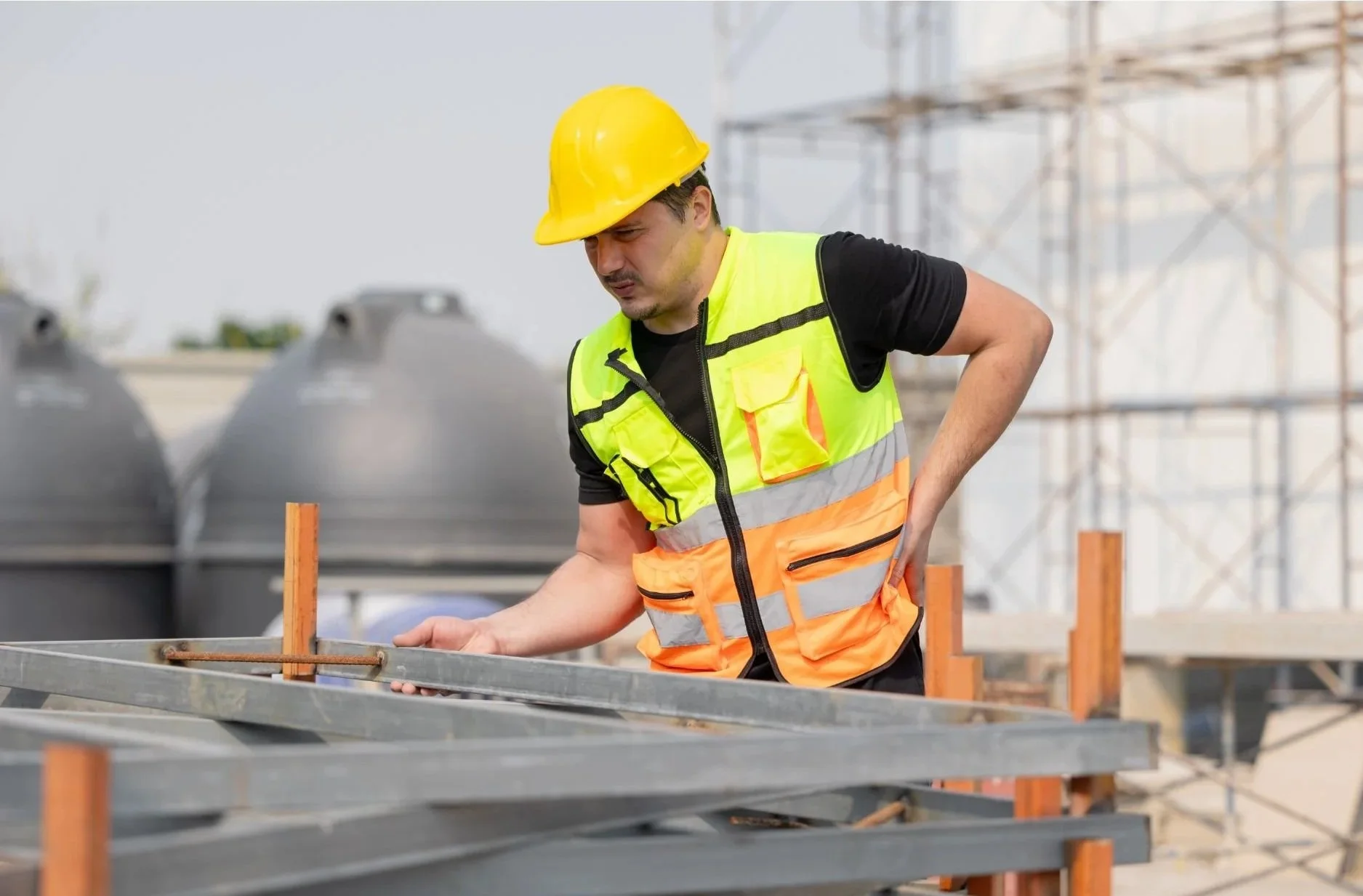 A construction worker in a yellow safety helmet and a neon orange safety vest inspects metal framing on a construction site.