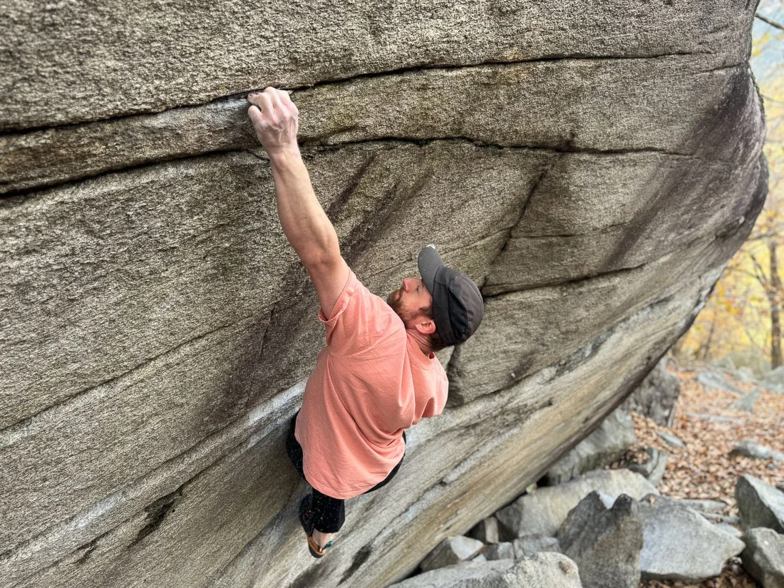 A person in a pink shirt and black shorts climbing a large overhanging rock in an outdoor setting with trees and rocks around.