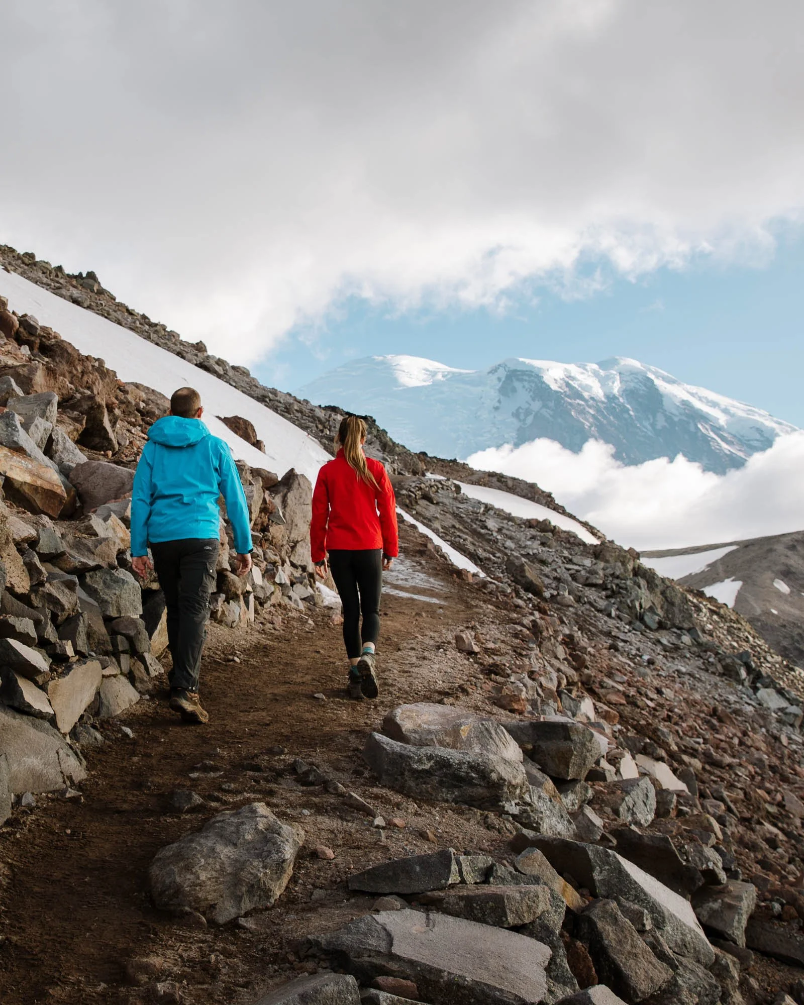 Two hikers, one in a blue jacket and one in a red jacket, walking on a rocky trail in a mountainous area with snow and clouds, and a snow-capped mountain in the background.