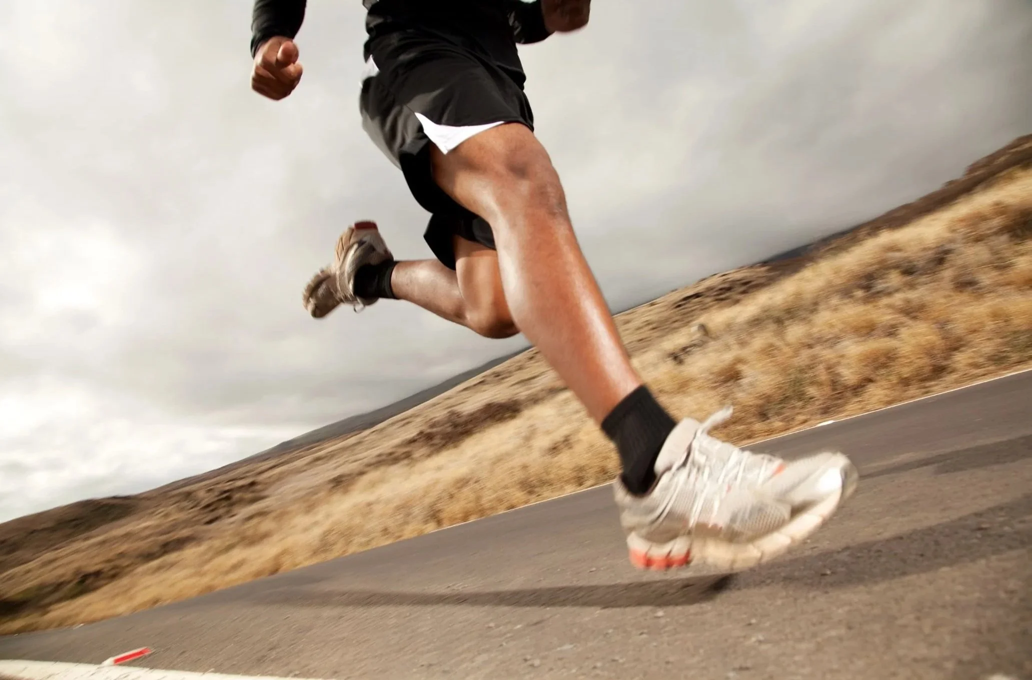 A person running on an outdoor paved trail in daylight with a hill and cloudy sky in the background.