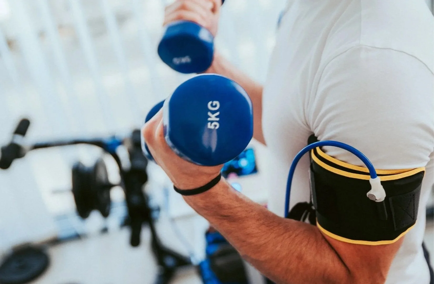 A person is lifting a 5-kilogram blue dumbbell in a gym, wearing a white shirt and a black armband with a yellow stripe. There are fitness bikes blurred in the background.