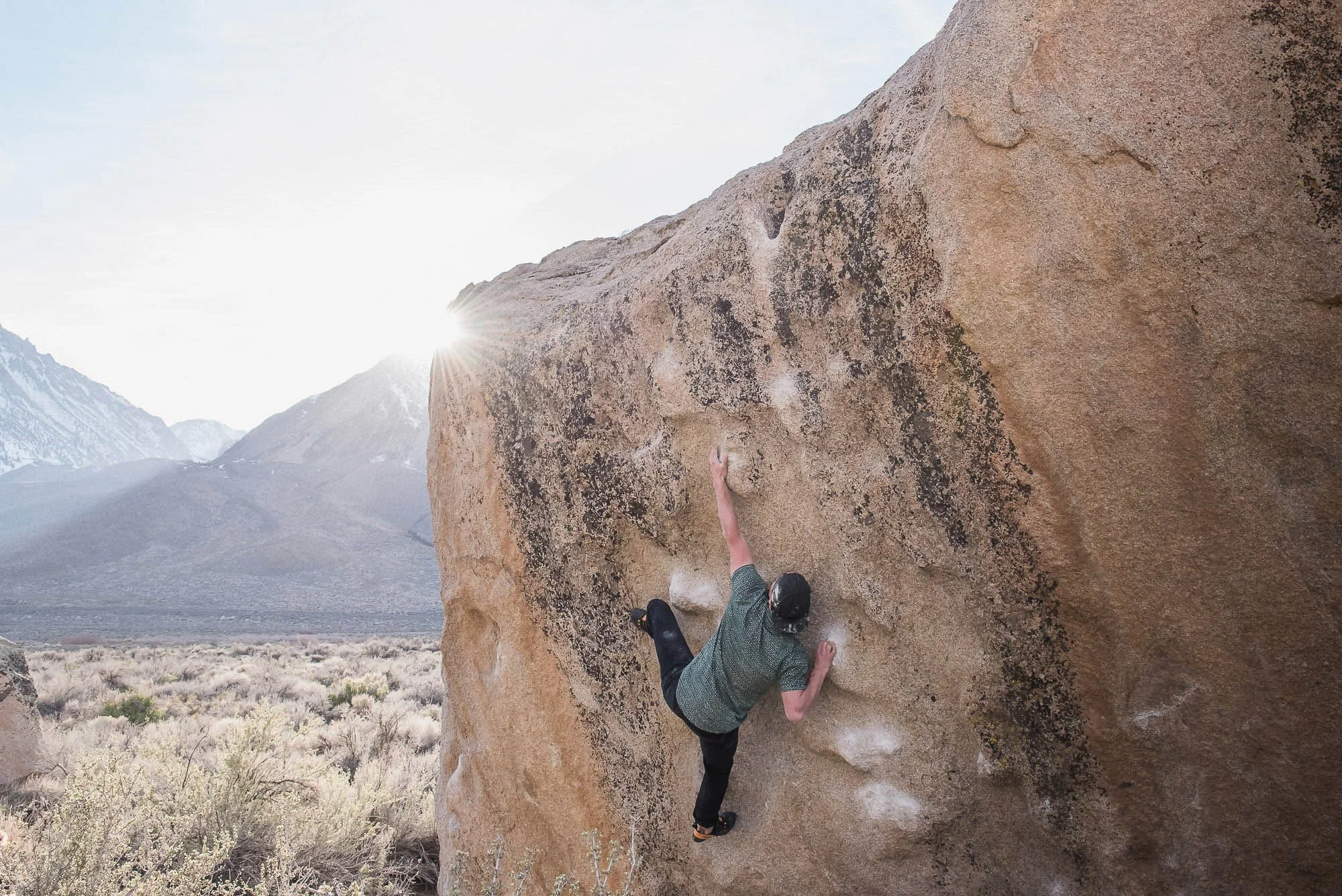 Person in a green shirt and black pants rock climbing on a large boulder outdoors in a desert landscape with mountains in the background and the sun shining.