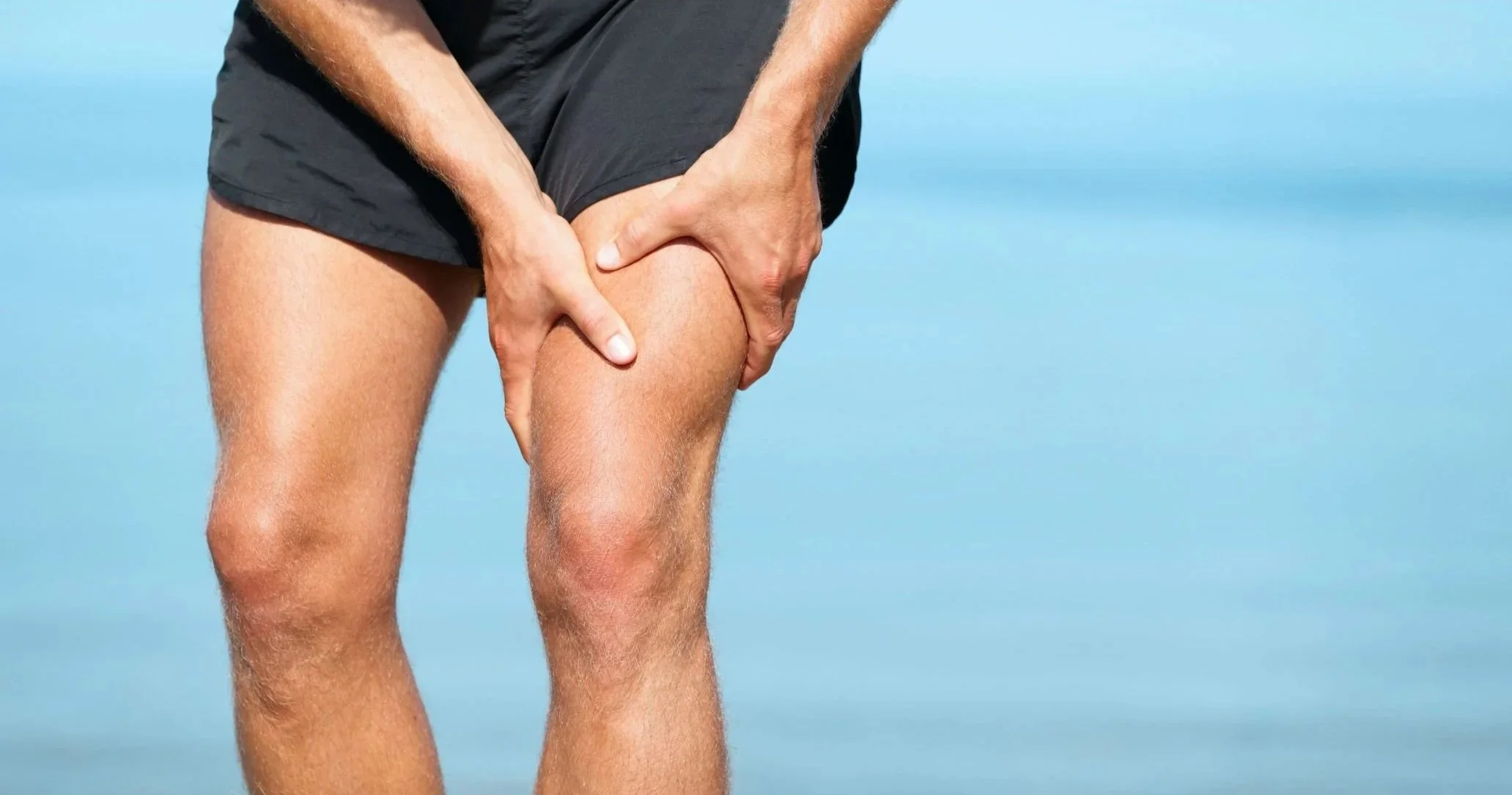 A person wearing black shorts on a beach, holding their injured right knee with their right hand, with an ocean and blue sky in the background.