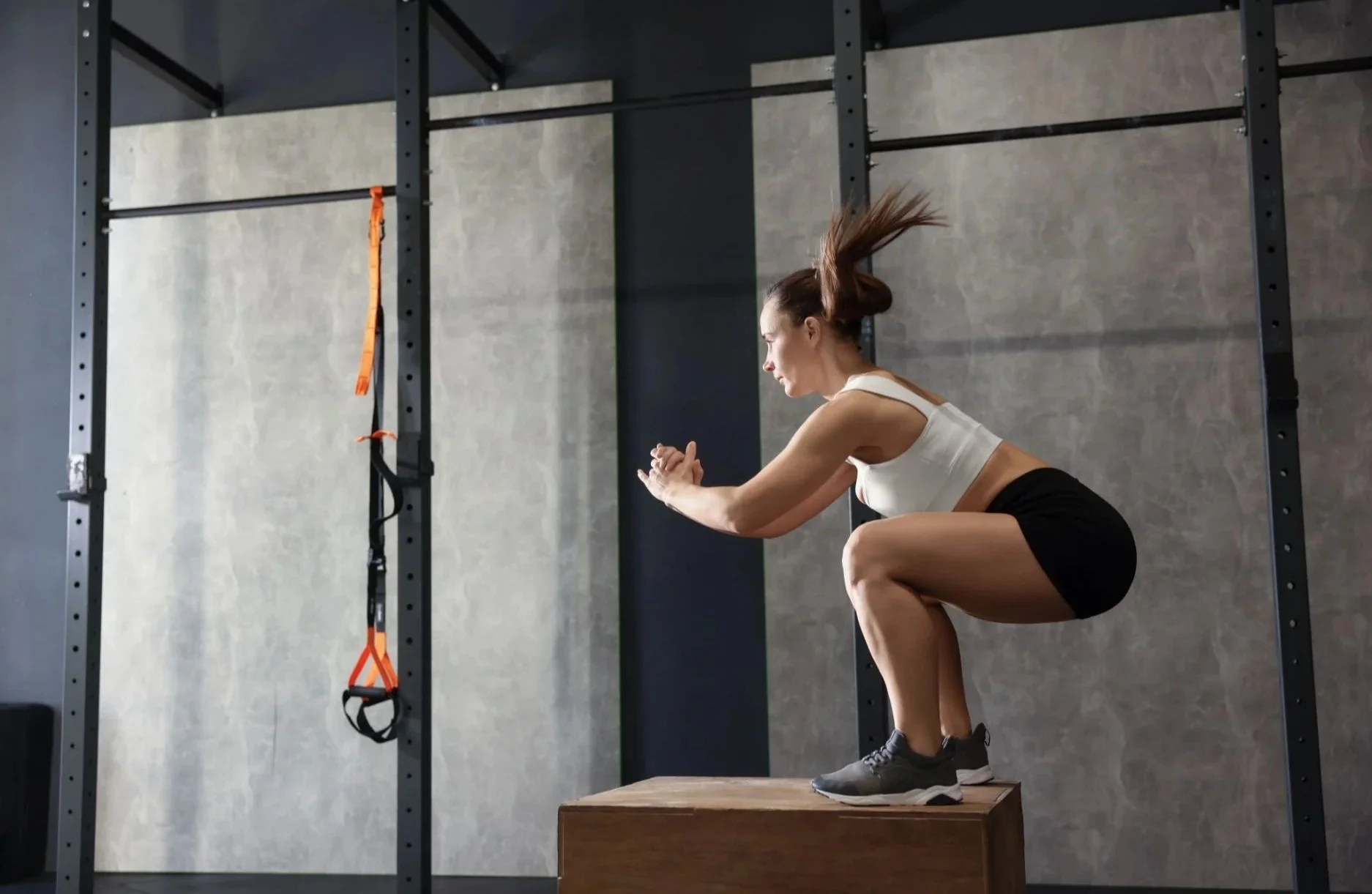 A woman in workout clothes squatting on a wooden box in a gym, with her hair swinging forward as she focuses on her exercise.