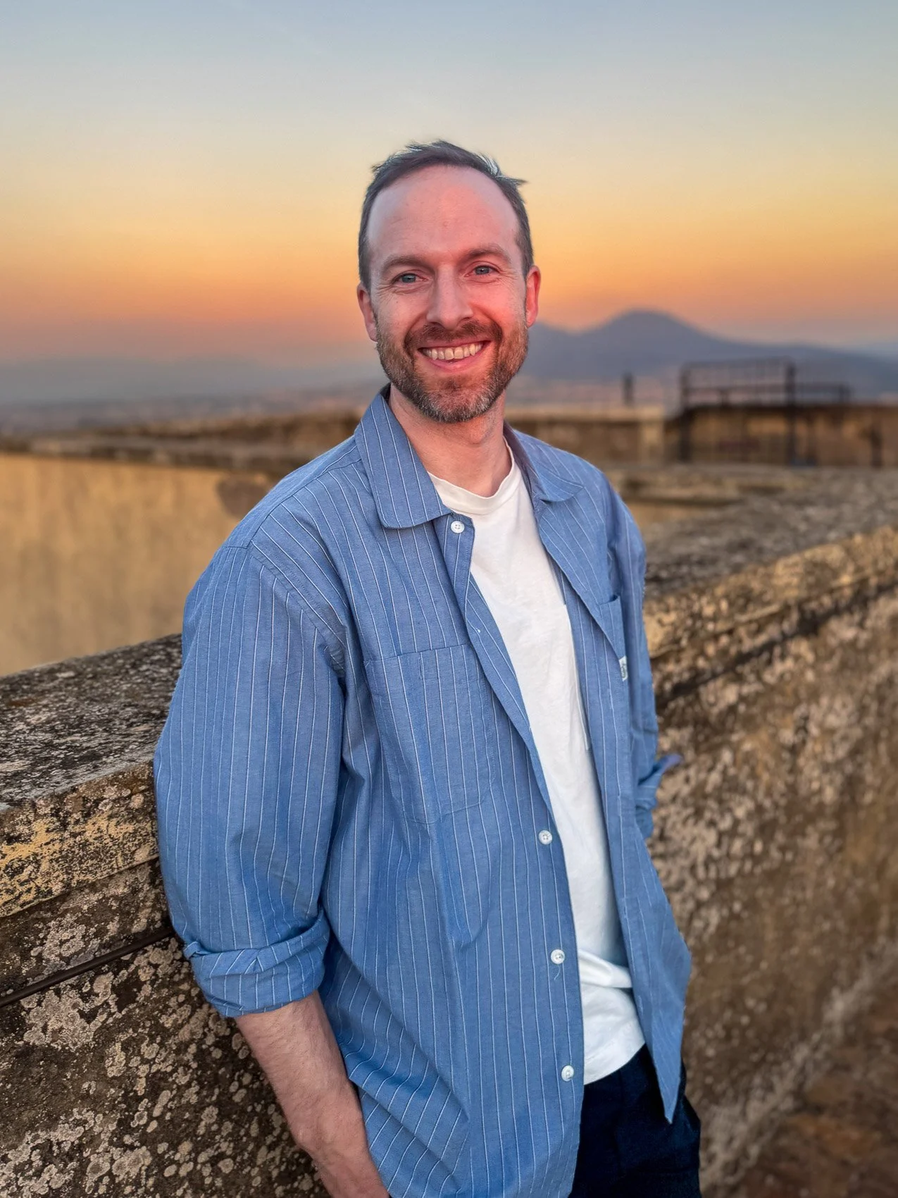 A smiling man with a beard and short hair in a blue striped shirt and white t-shirt standing outdoors by a stone wall during sunset with hills in the background.