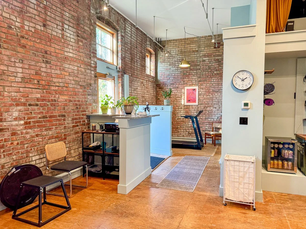 Interior of a loft-style apartment with exposed brick walls, hardwood floors, a treadmill, a modern counter with plants, and a bookshelf with exercise equipment.