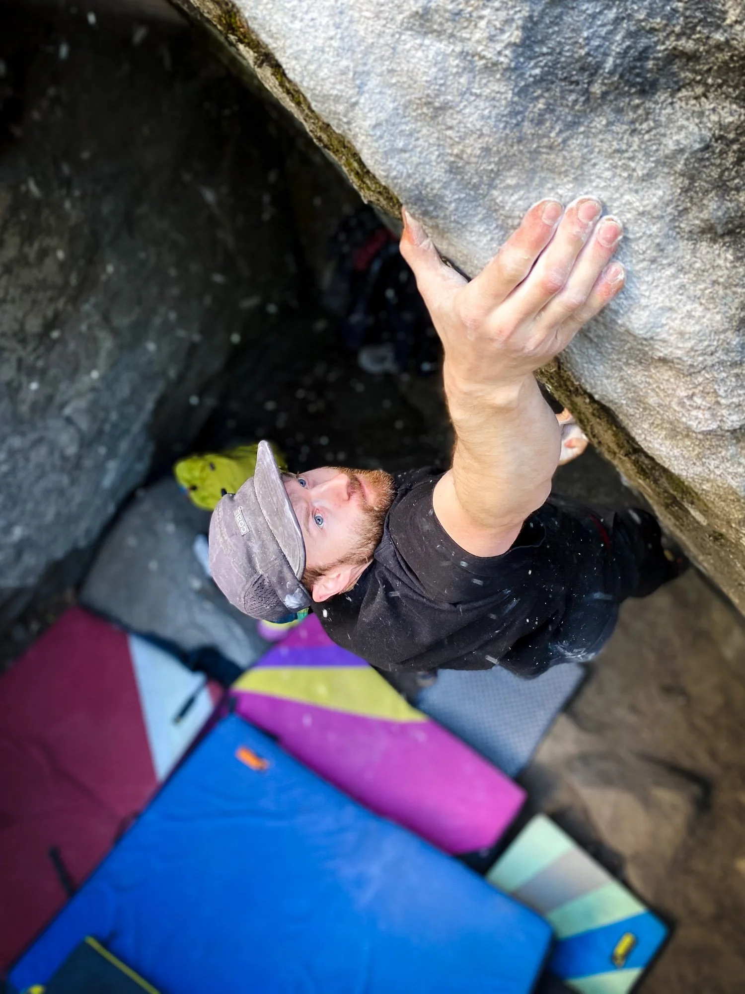 A man rock climbing outdoors, reaching for a handhold on a steep rock face with safety equipment and a mat below.