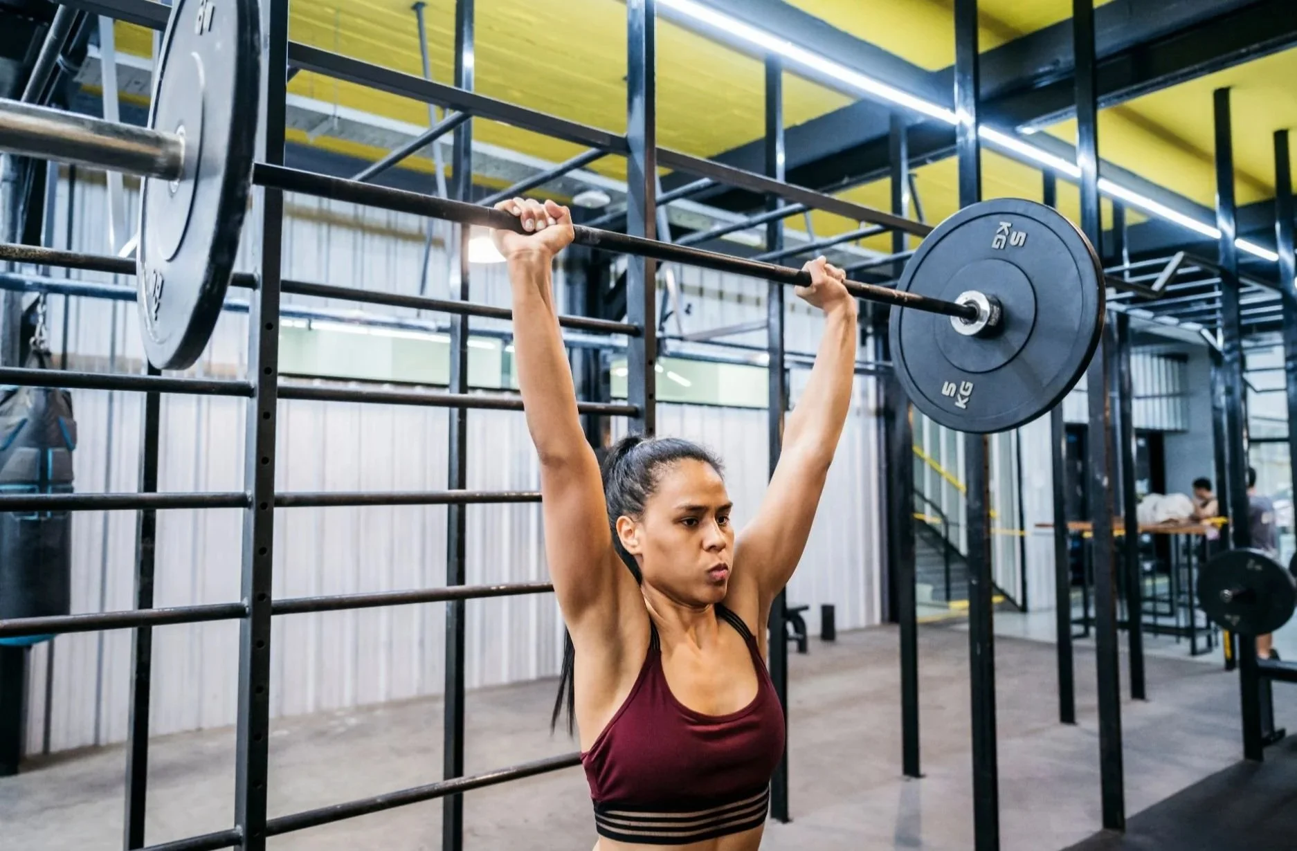 A woman in a gym lifting a barbell with weights overhead.