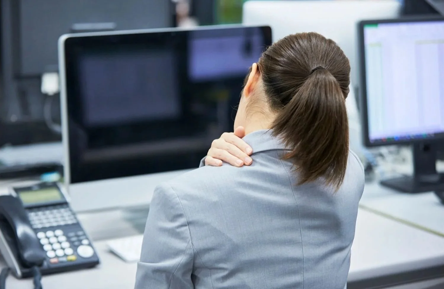 Back of a woman with brown hair in a ponytail, wearing a gray blazer, experiencing neck pain at her office desk, with multiple computer monitors and office equipment around.