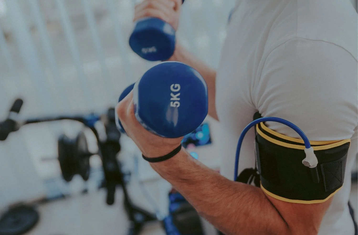 A person in a white shirt lifting a 5kg blue dumbbell in a gym, with exercise equipment visible in the background.