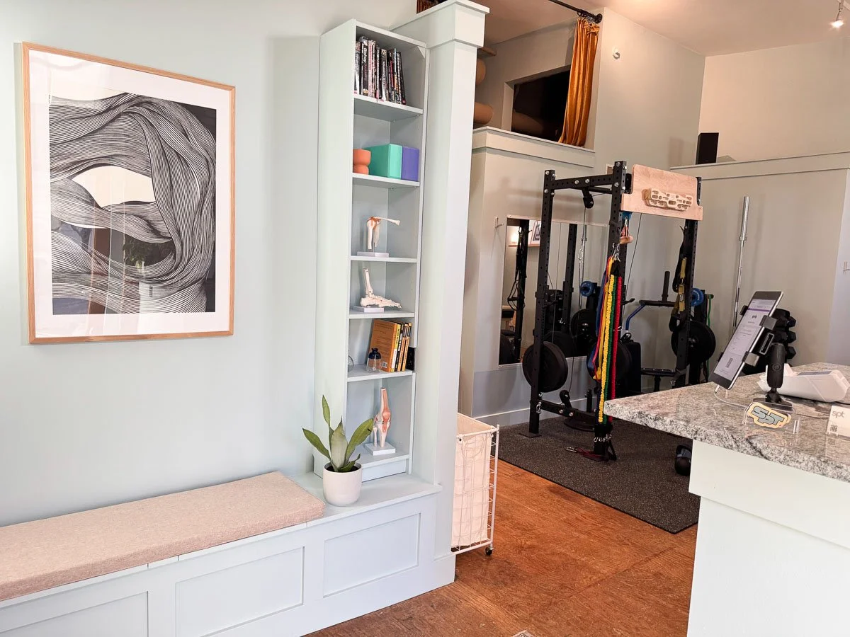 Interior of a home gym with workout equipment, a granite kitchen counter, a wall-mounted artwork, and a built-in shelf with fitness models and books.