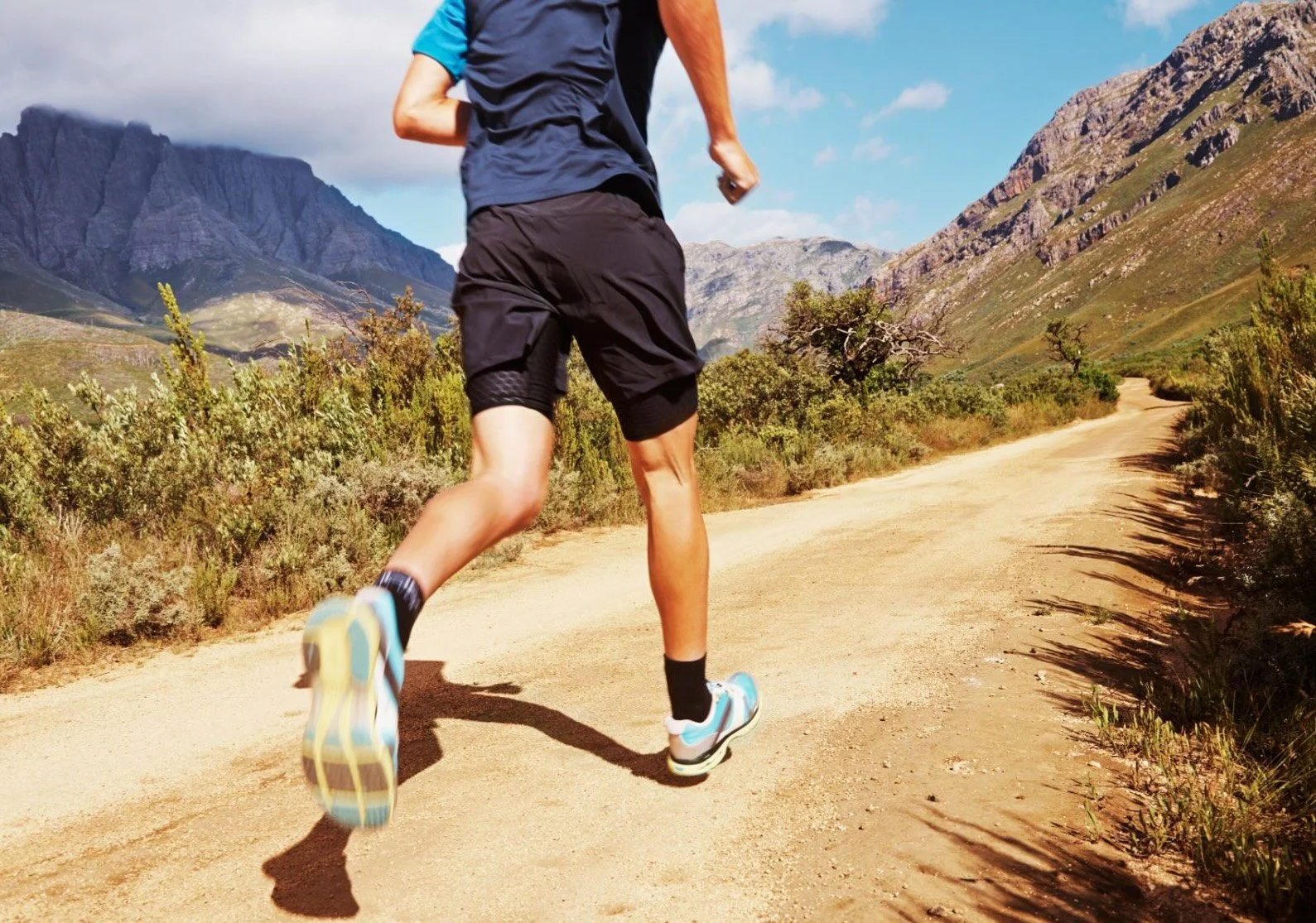 A person running on a dirt trail through a mountainous landscape with green shrubs, rugged mountains, and a partly cloudy sky.