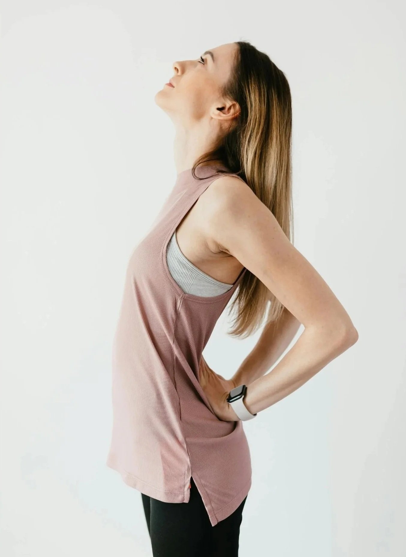 A woman standing sideways, stretching her back with her hands on her hips, wearing a sleeveless pink top and a smartwatch on her wrist.