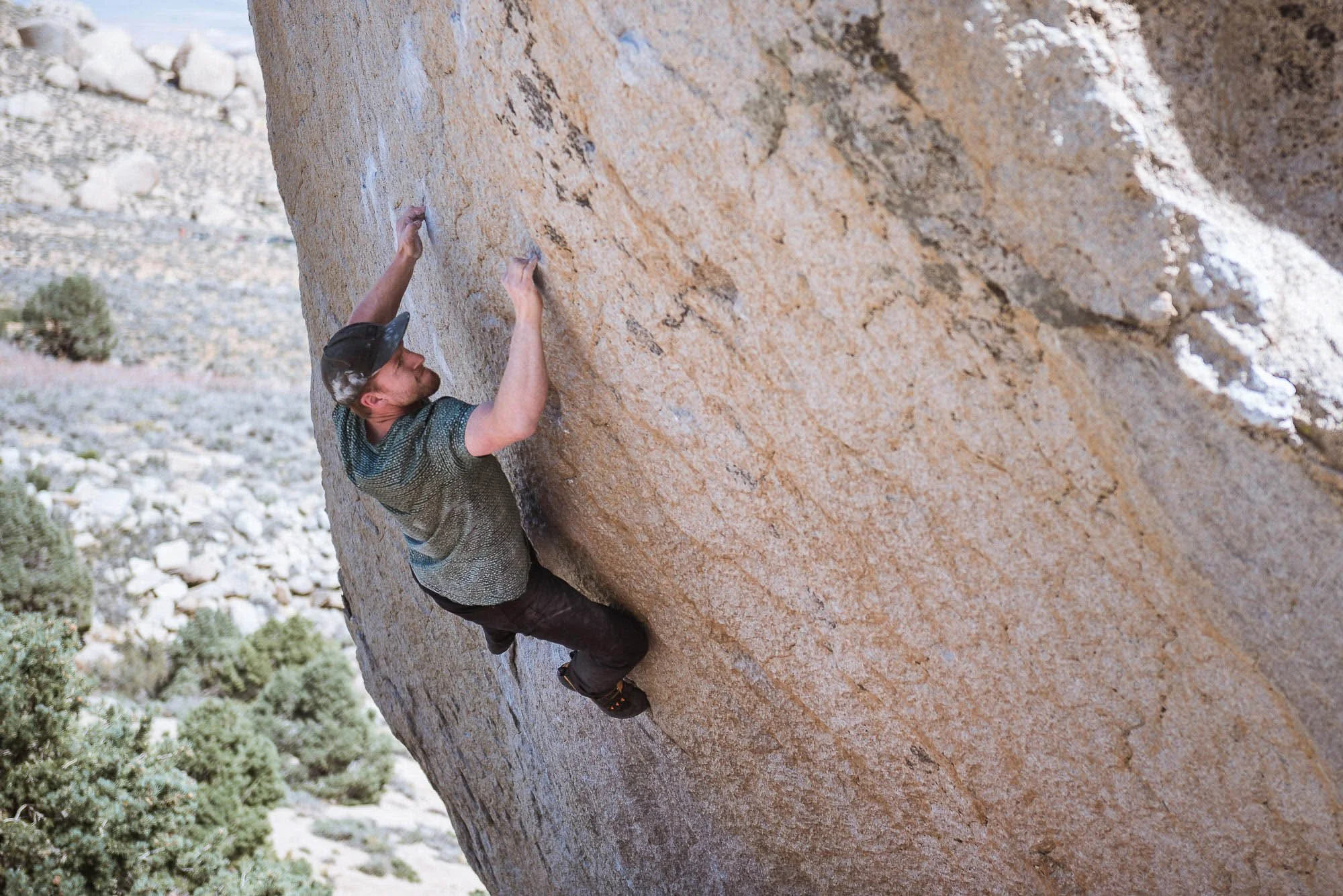 A man in a gray shirt and black hat rock climbing on a large, sloped, tan granite boulder outdoors.