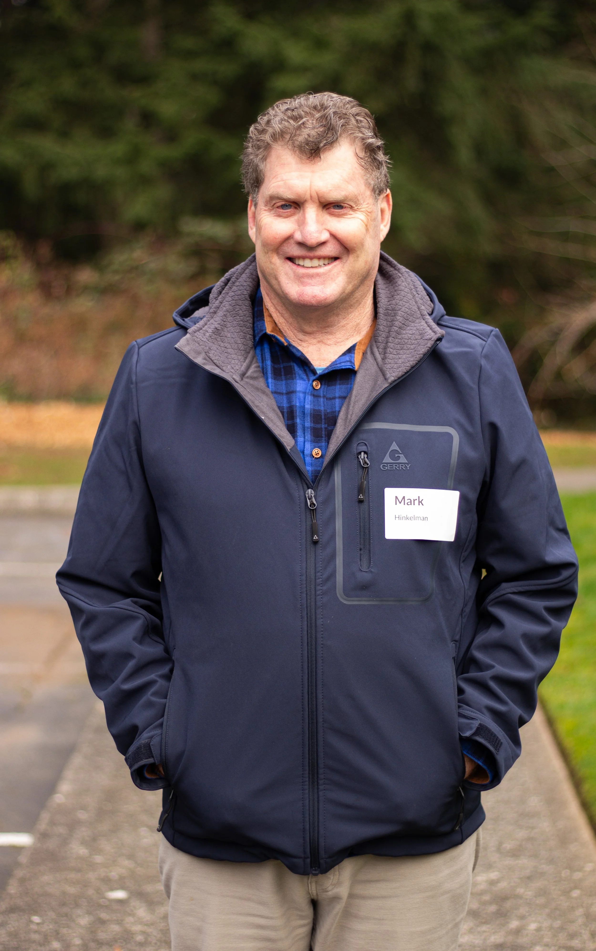 A smiling man outdoors wearing a dark blue jacket and a blue plaid shirt.