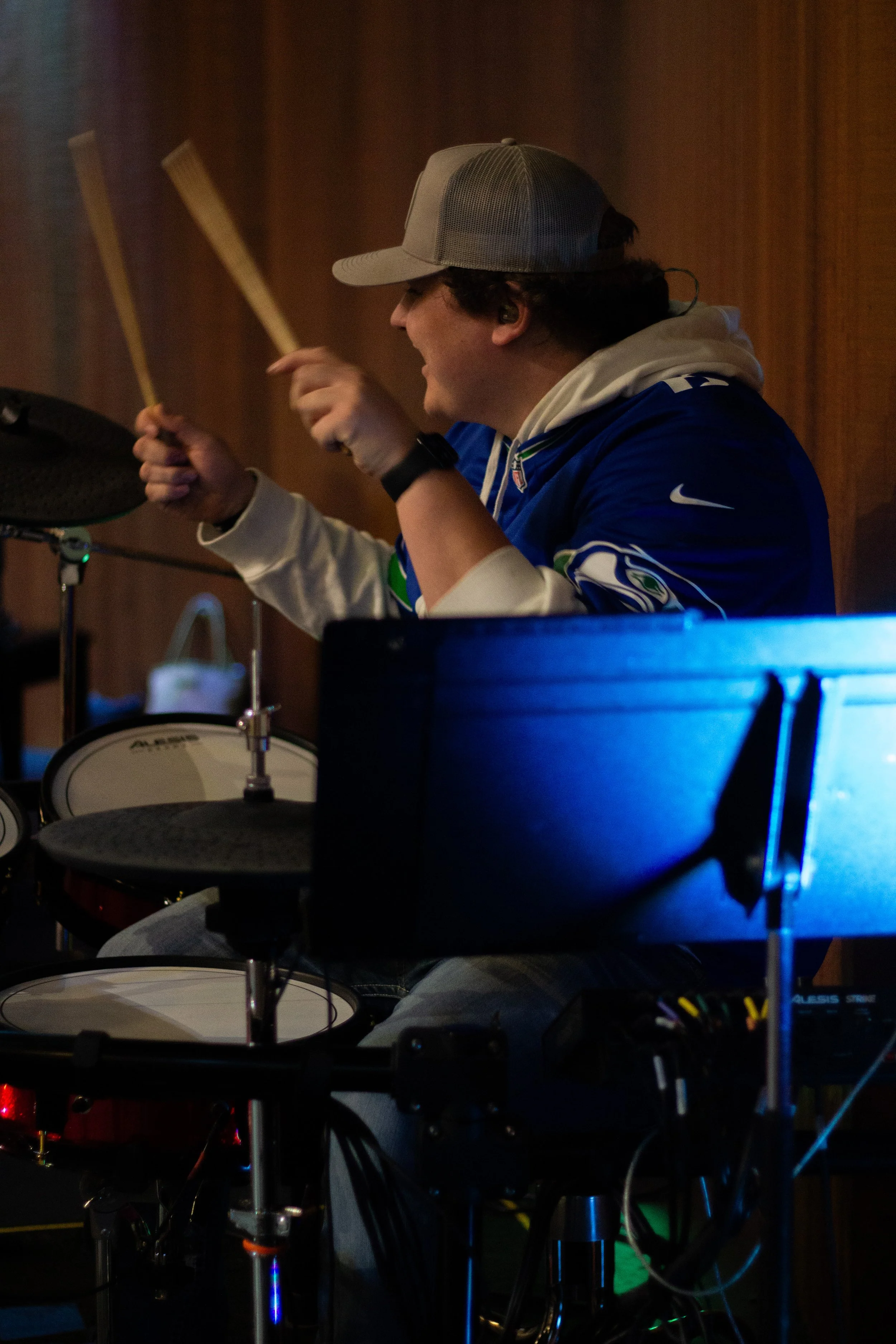 A man playing electronic drums, wearing a gray baseball cap, blue and white Seattle Seahawks jacket, and white hoodie, in a dimly lit room.