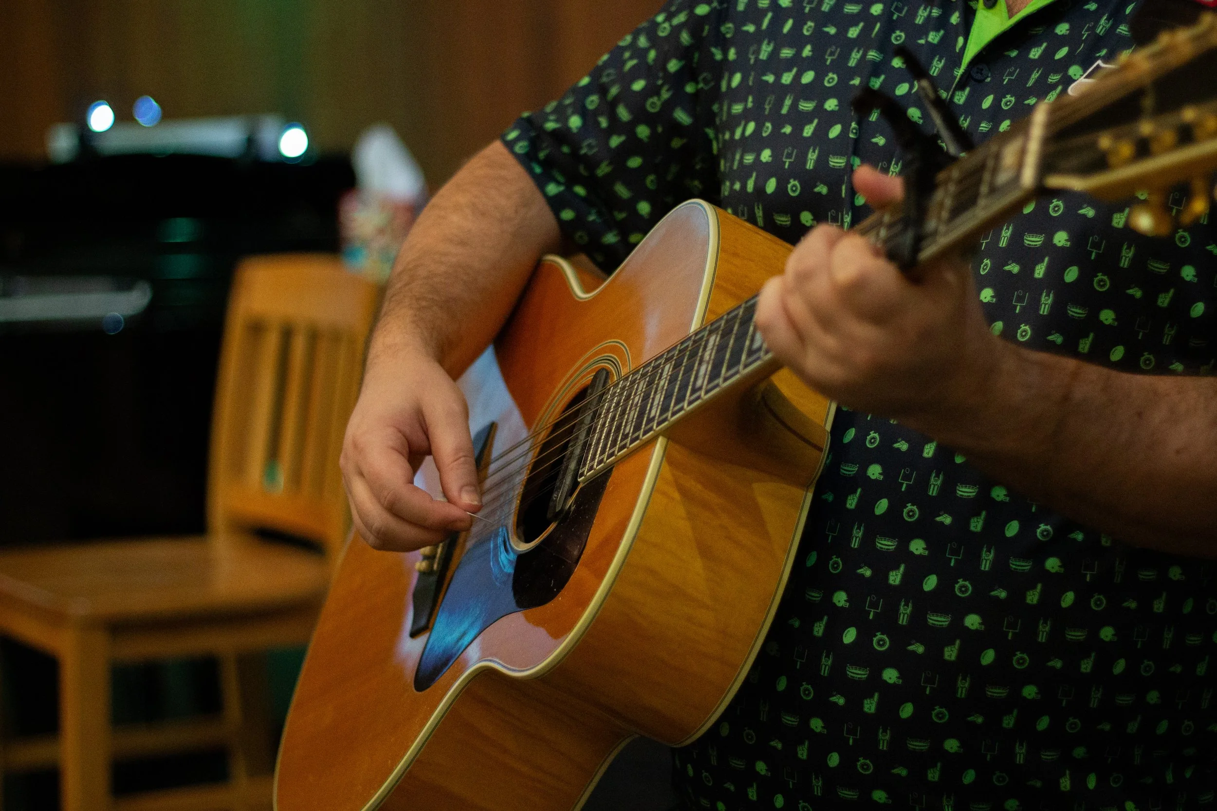 Person playing an acoustic guitar, wearing a black shirt with green musical notes pattern, in a room with wooden chairs and a TV in the background.