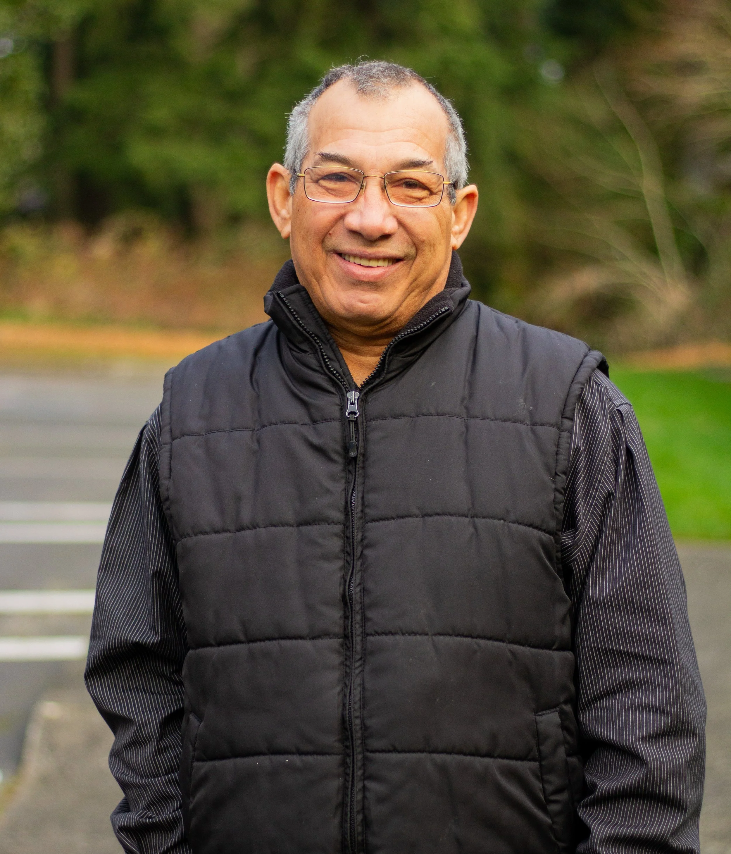 A smiling man with glasses outdoors, wearing a black padded vest over a long-sleeve shirt with vertical stripes, standing on a sidewalk with blurred greenery in the background.