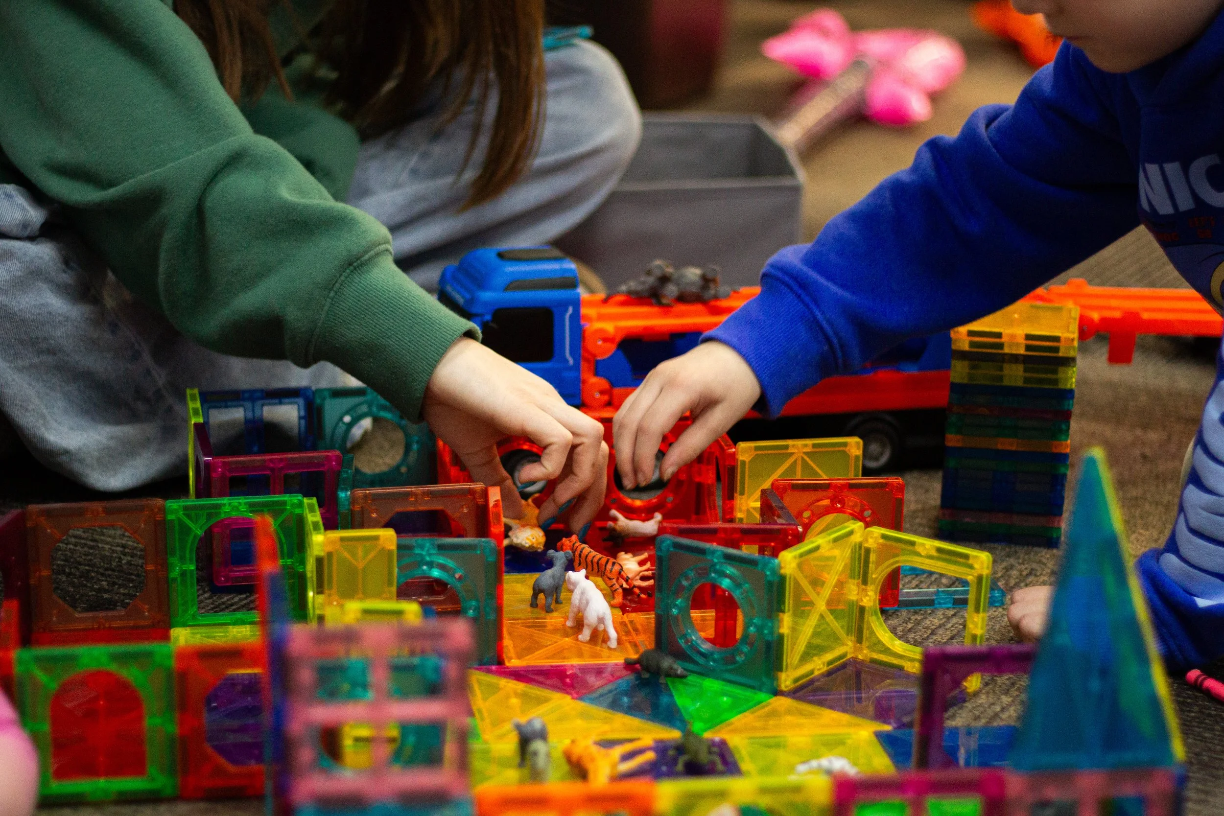 Two children playing with colorful magnetic building tiles and small animal figurines on a brown carpeted floor.