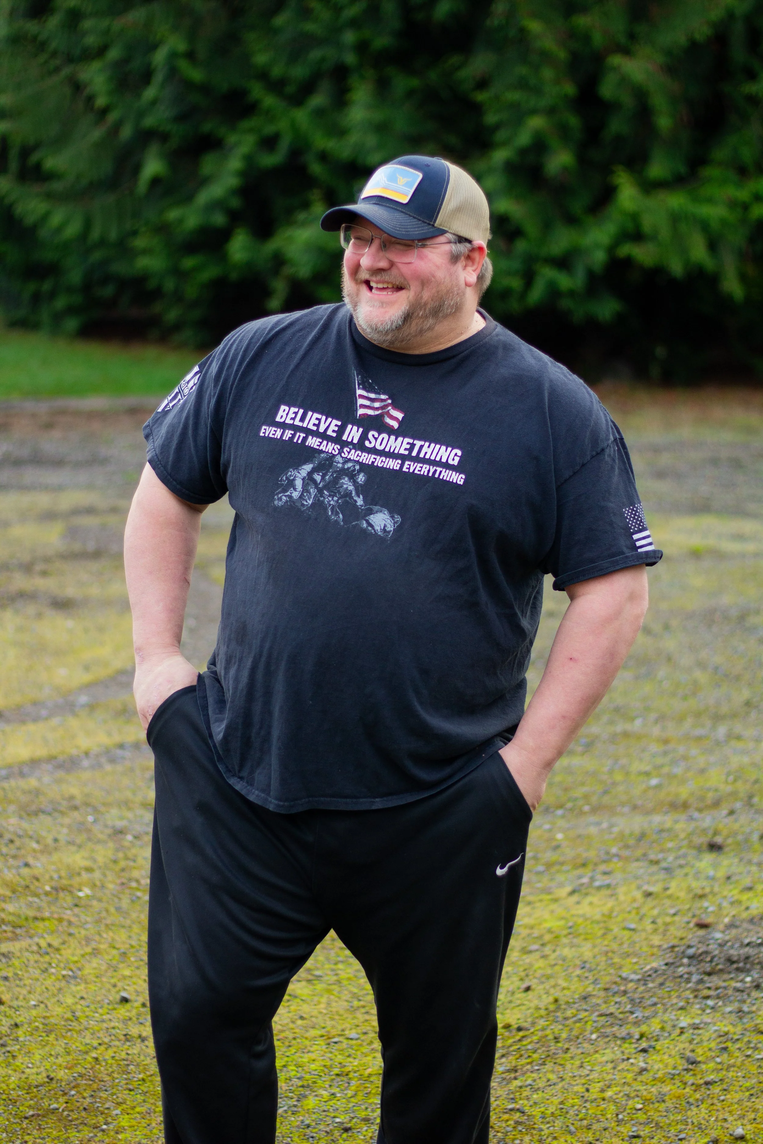 A man with glasses and a beard wearing a black t-shirt, black pants, and a baseball cap, standing outdoors with his hands in his pockets, smiling. The background features green trees and mossy ground.