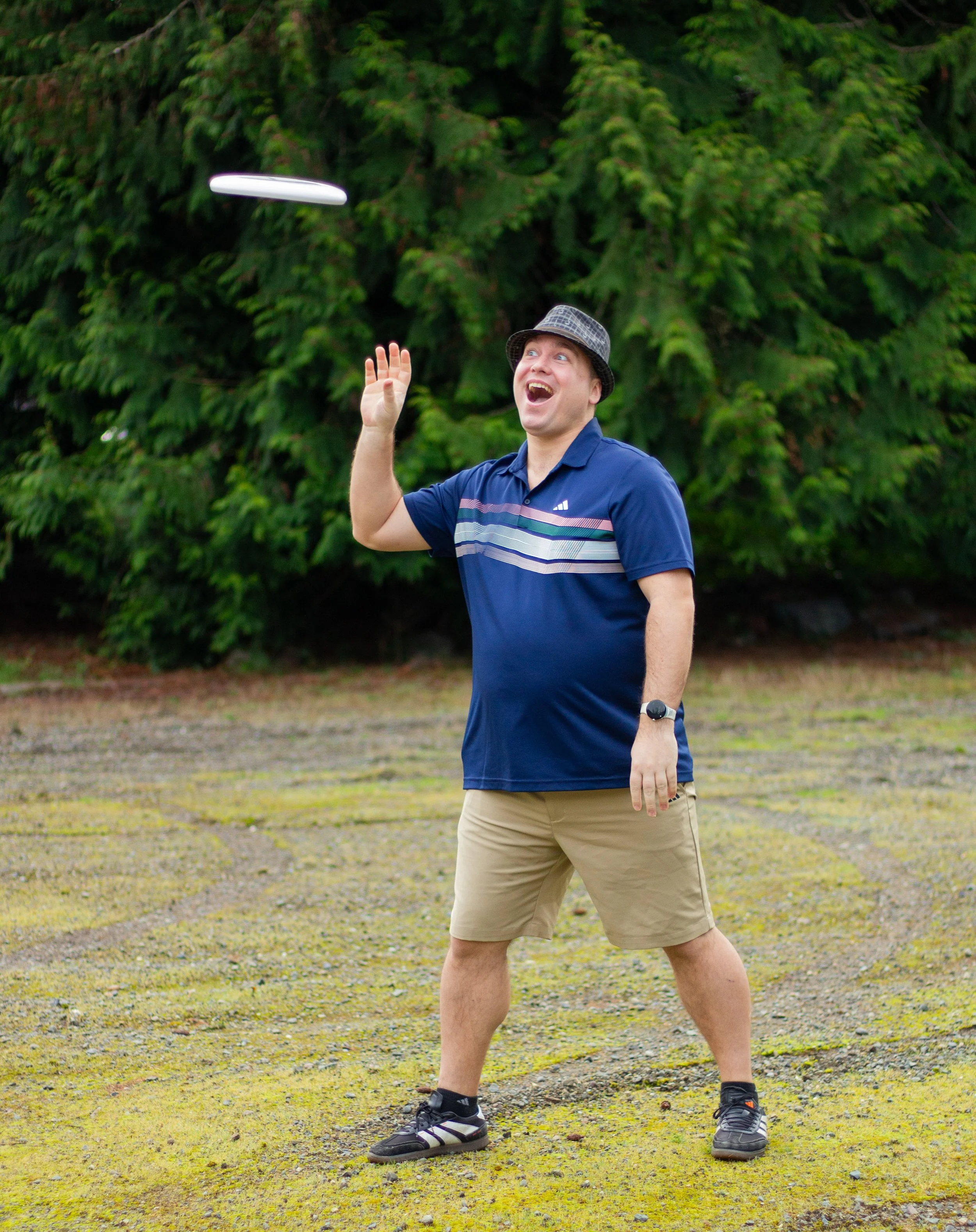 A man wearing a hat, navy blue polo shirt, and khaki shorts playing frisbee outdoors near bushes.