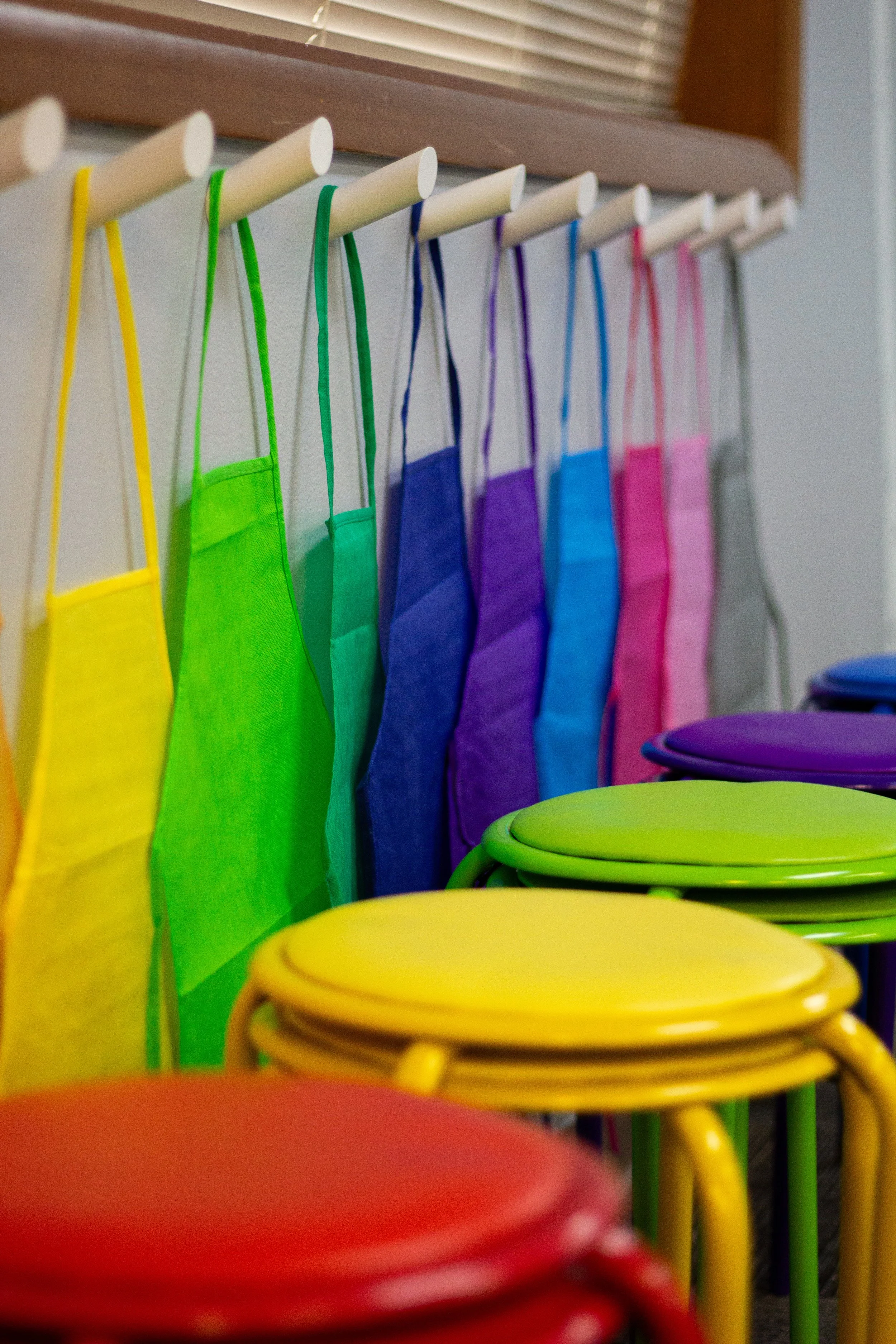 Colorful fabric masks hanging on a wall hook with brightly colored chairs stacked below.