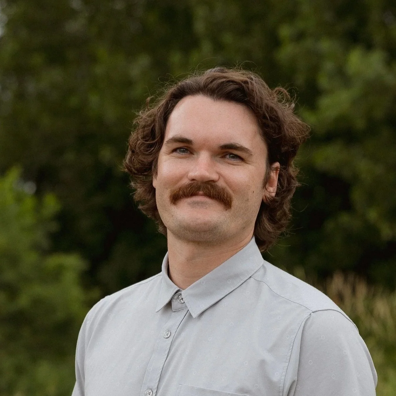 Portrait of a smiling man with curly brown hair and a mustache wearing a light gray button-down shirt outdoors