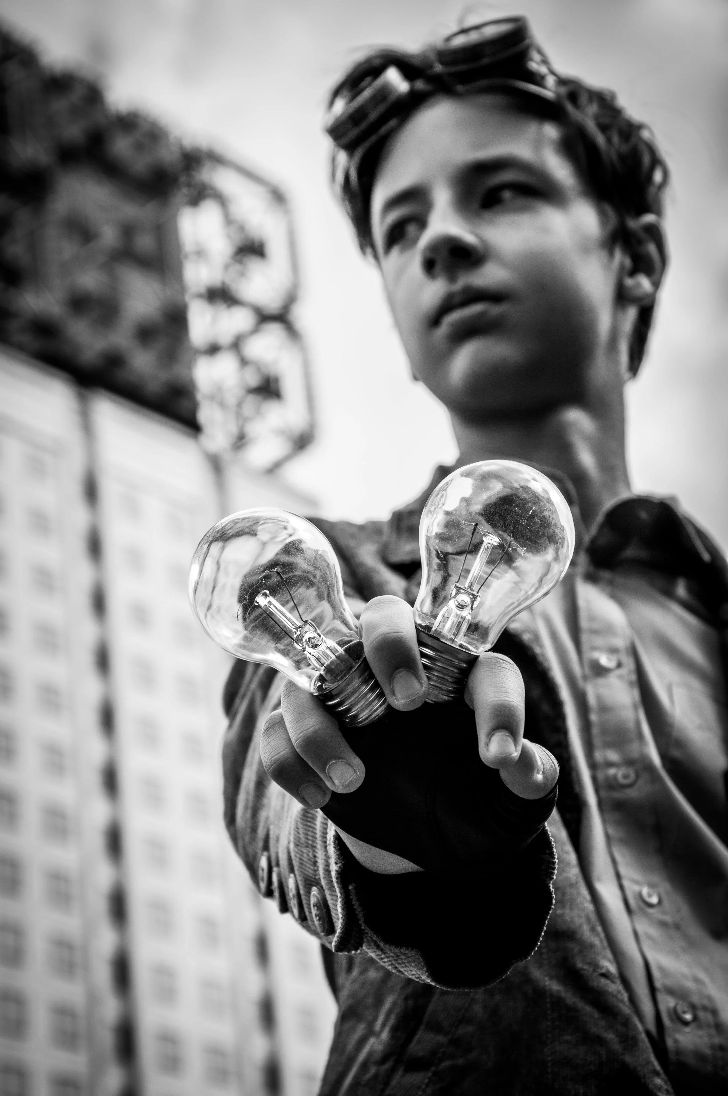 A young person wearing goggles on their head is holding three clear incandescent light bulbs towards the camera. The background includes a blurred billboard and a brick wall.