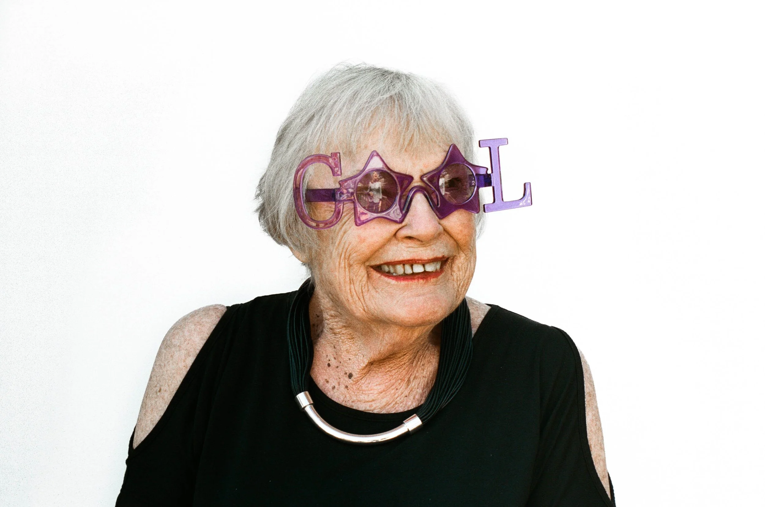 An elderly woman wearing fun purple glasses that spell out 'GOLD'. She is smiling and has short gray hair, wearing a black top with a cut-out shoulder design, a silver necklace, and against a plain white background.