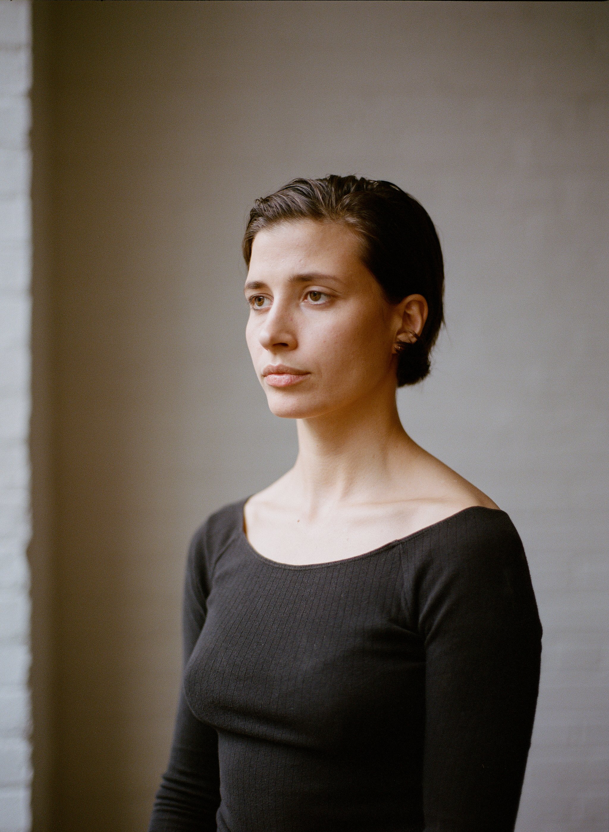 A woman with short dark hair wearing a black long-sleeve top, standing indoors against a neutral background, looking thoughtfully to the left.