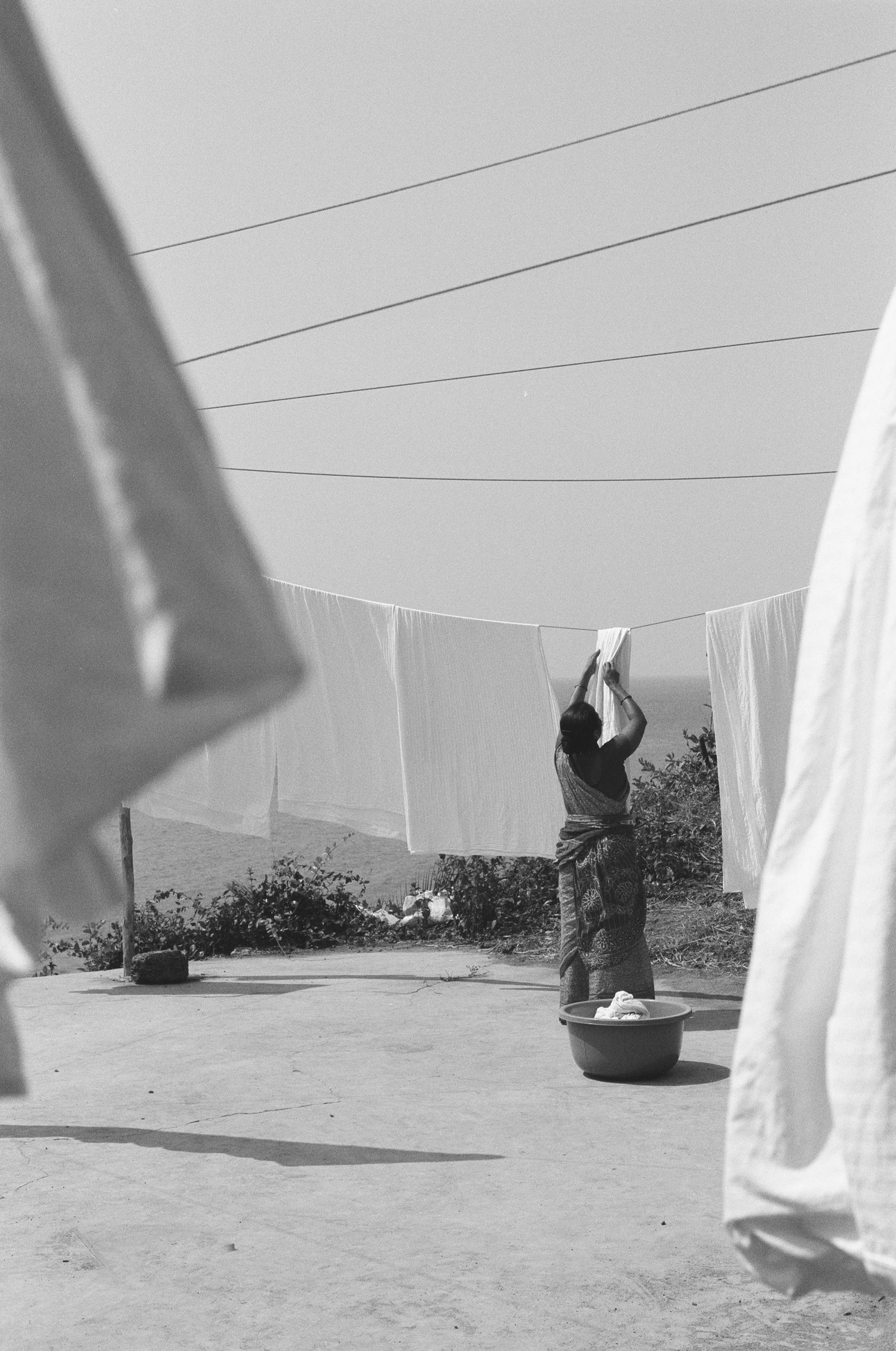 A woman hanging laundry on a clothesline outdoors, with plants and a distant landscape in the background.