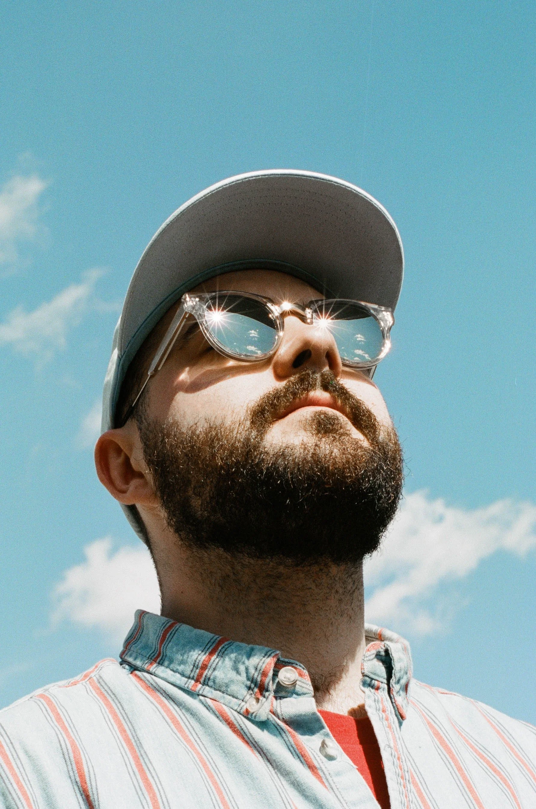 A man with a beard and sunglasses looking up at the sky, wearing a baseball cap and a striped button-up shirt.