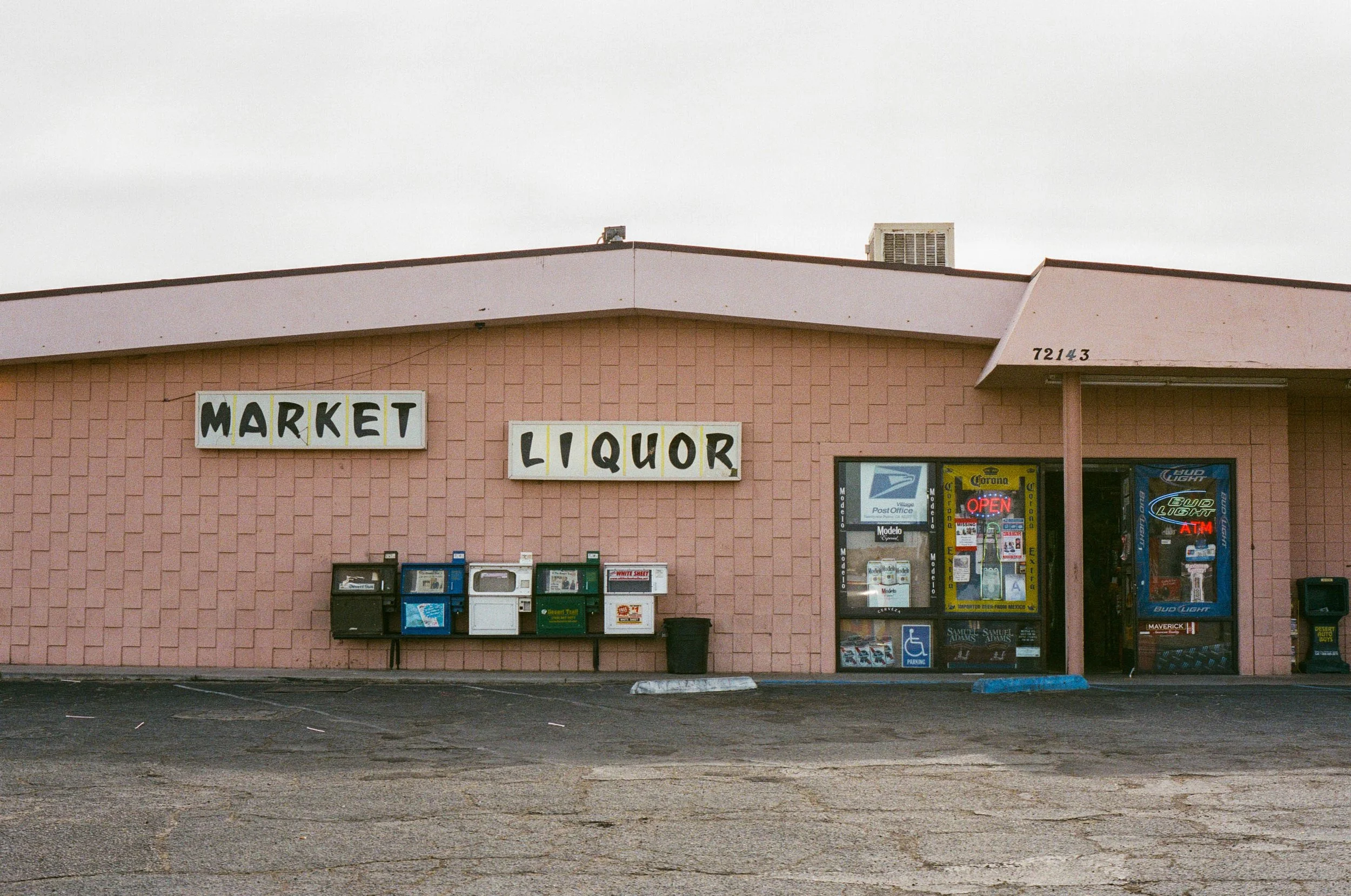 A single-story pink building with signs that say "Market" and "Liquor." The building has a small parking lot in front with an uneven asphalt surface. There are newspaper vending machines and a trash can outside, and a glass door entrance with various signs and a neon "Open" sign. The building's address is 7214.