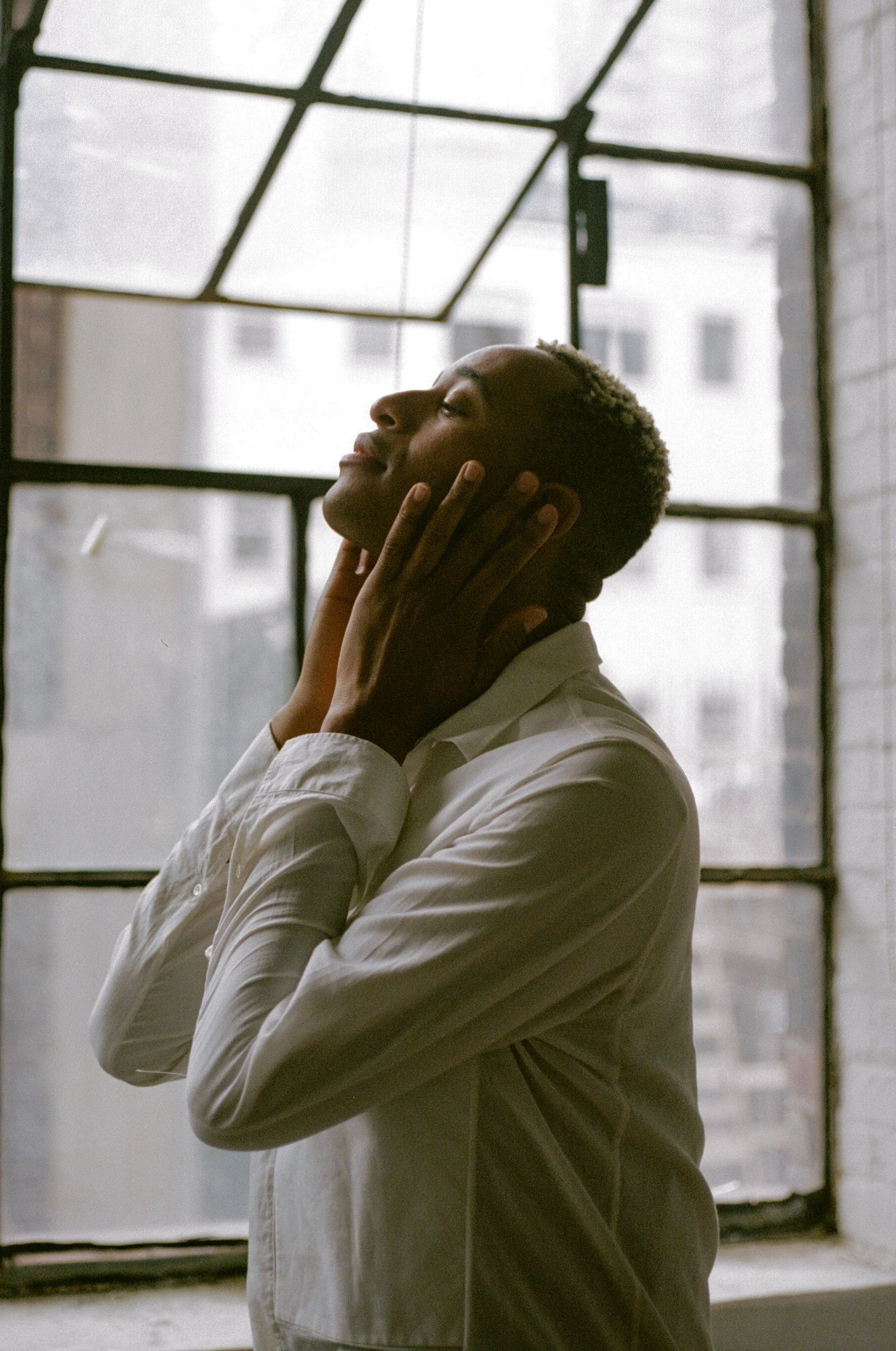 A person in a white shirt standing by a large window, head tilted upwards, touching their face with both hands, with a thoughtful expression.